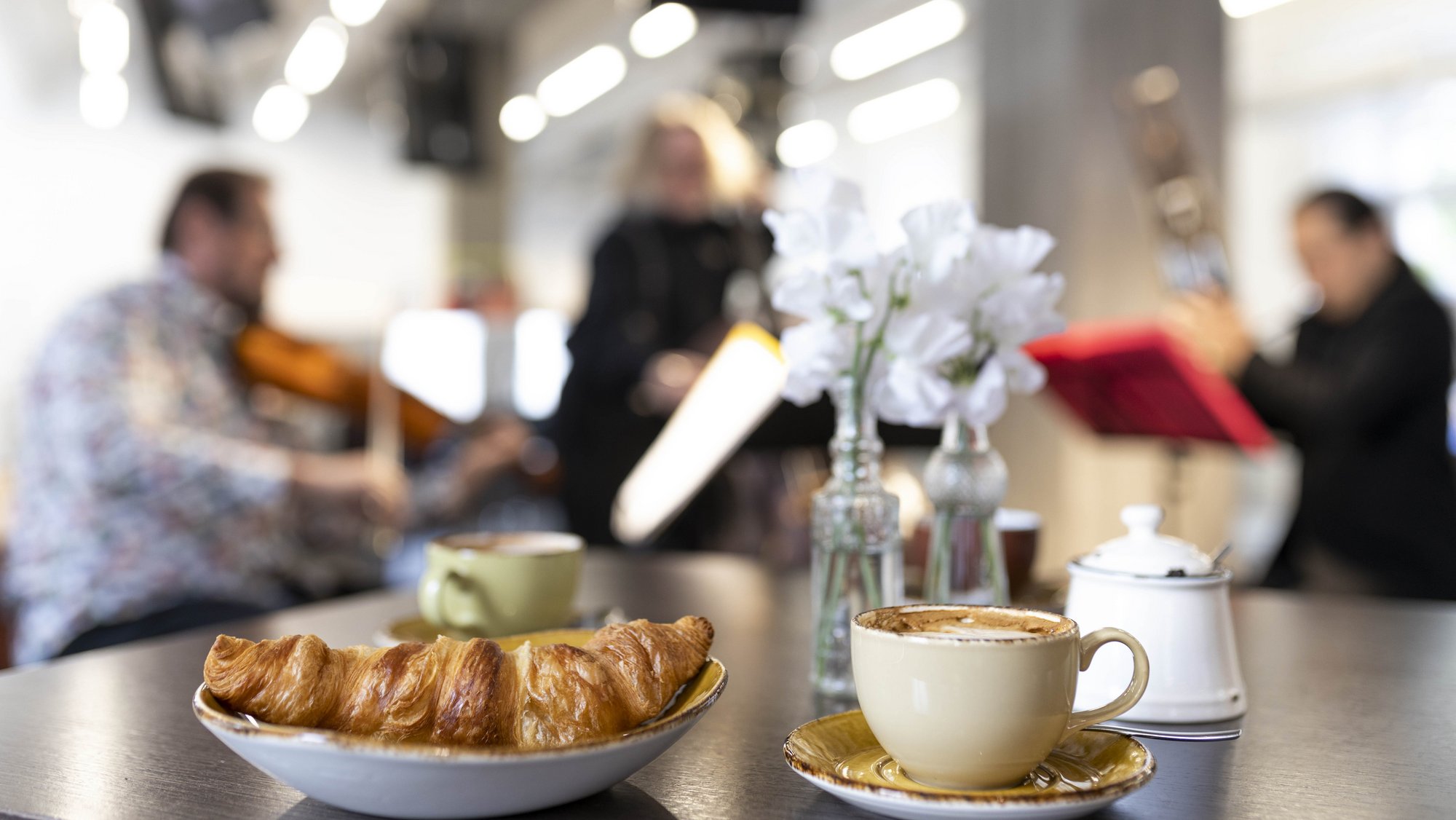 Croissant and coffee on a table, musicians play in the background