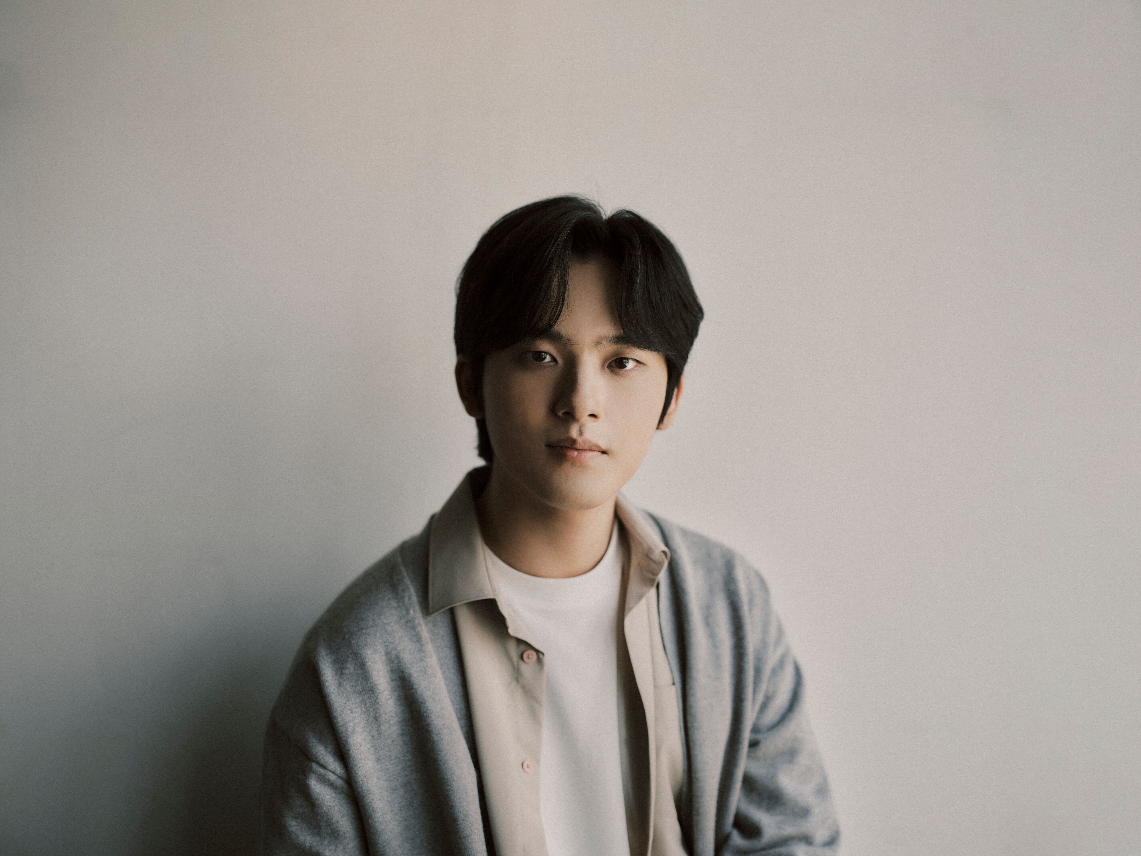 Yunchan Lim mit mittellangen dunklen Haaren, die ein weißes Hemd und eine graue Strickjacke trägt, sitzt vor einem schlichten hellen Hintergrund und schaut ruhig in die Kamera.