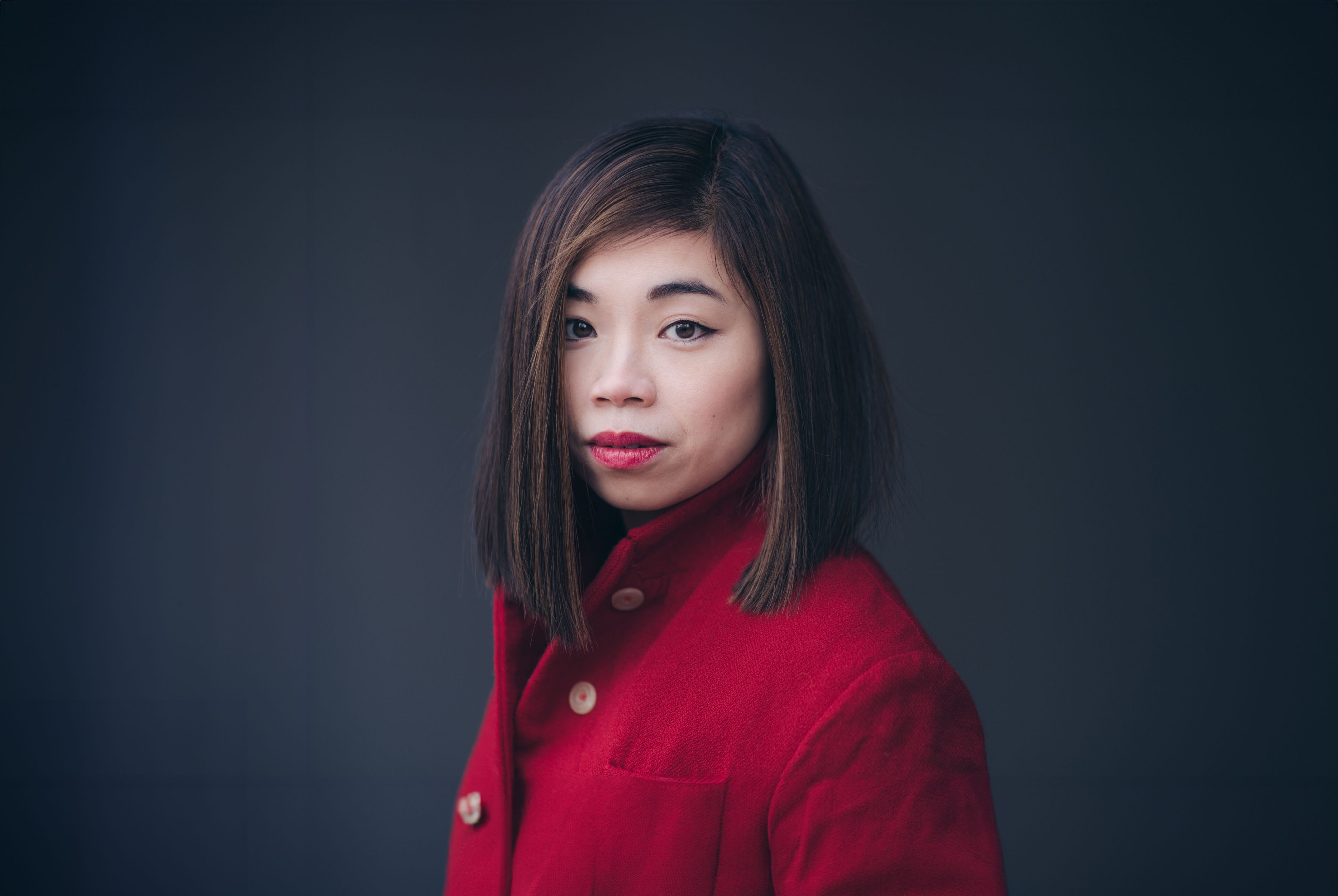 Elim Chan mit glattem, schulterlangem dunklem Haar und rotem Lippenstift trägt einen roten Mantel und blickt vor einem schlichten dunklen Hintergrund direkt in die Kamera.