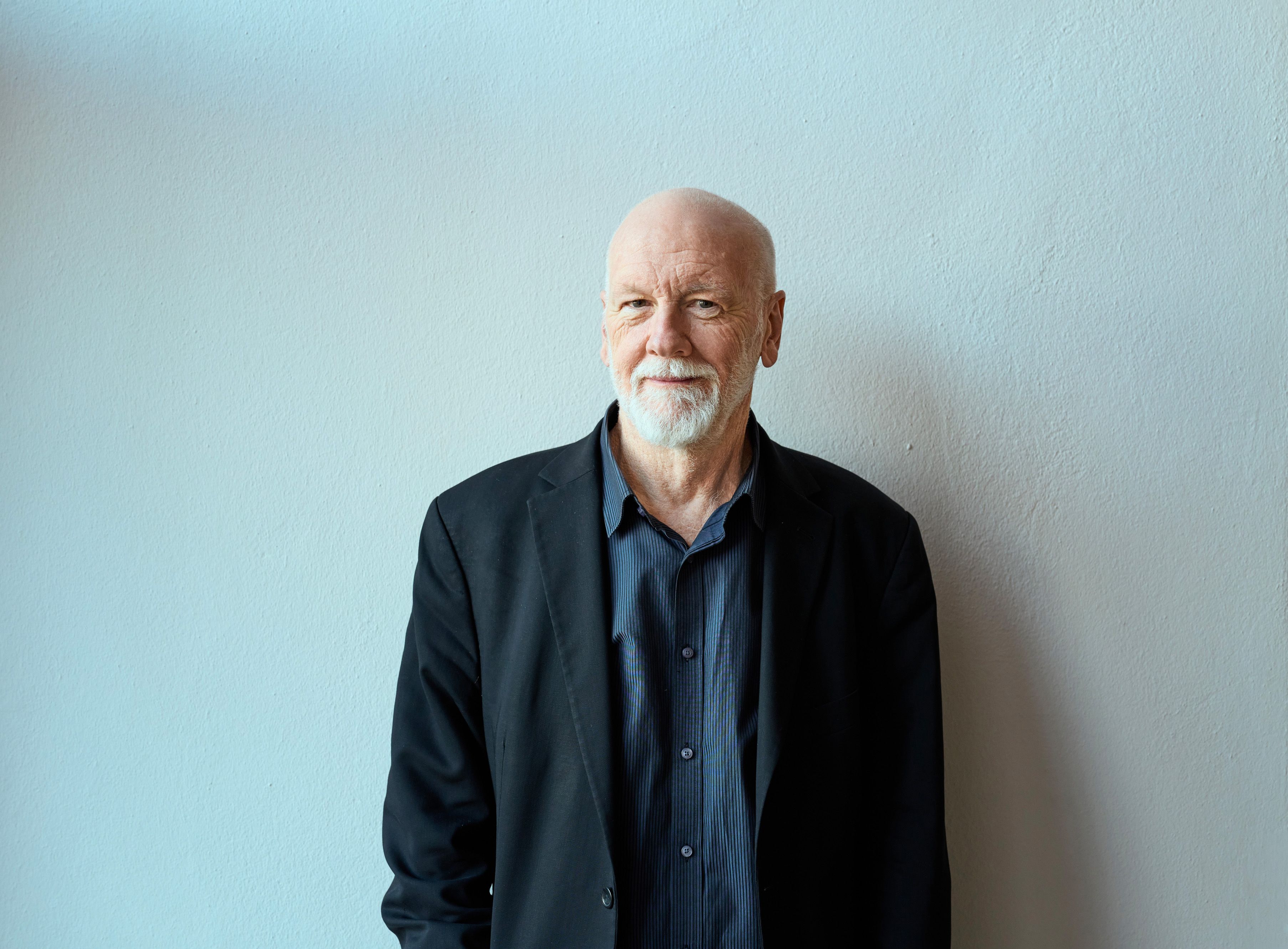 Ein älterer Mann mit Glatze, weißem Bart und Schnurrbart steht vor einer hellblauen Wand, trägt eine schwarze Jacke über einem dunklen Hemd und schaut mit einem leichten Lächeln in die Kamera.