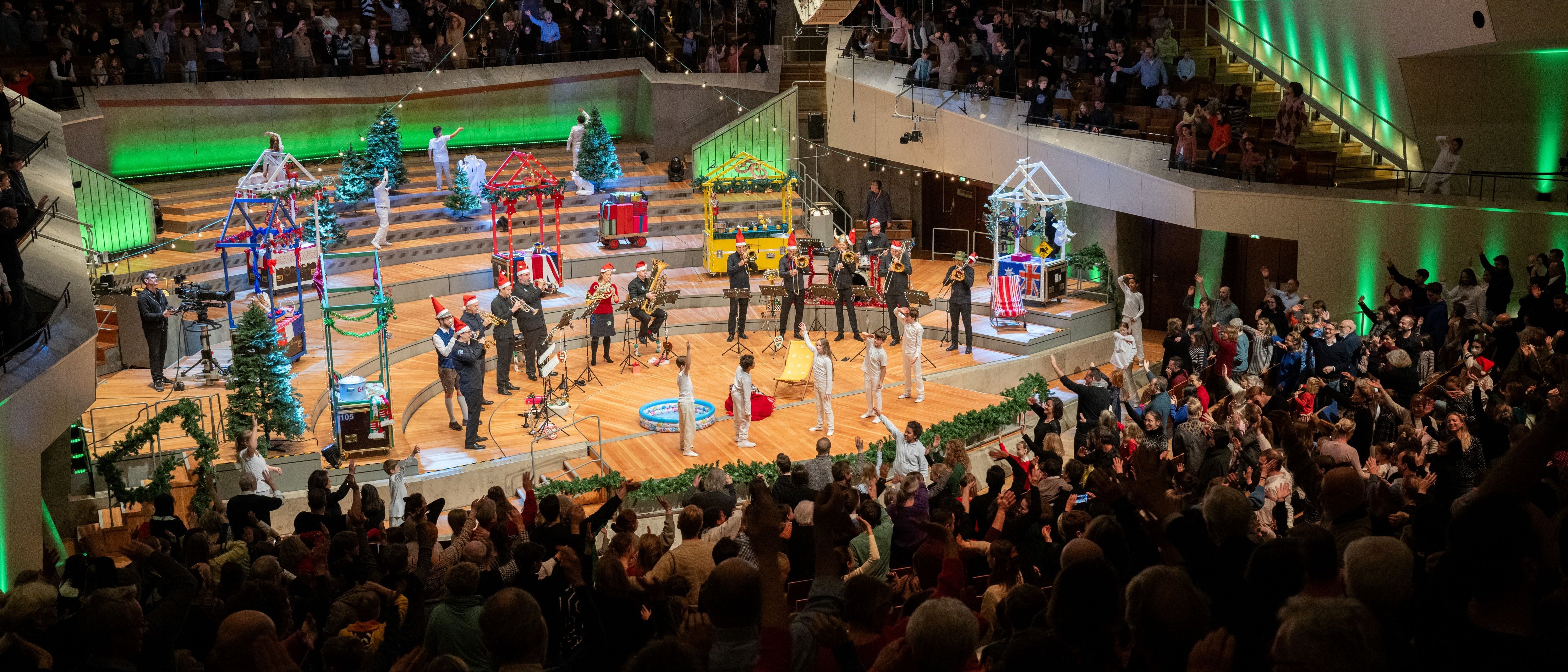 Ein lebendiger Konzertsaal mit weihnachtlicher Dekoration auf der Bühne. Das Publikum, das in geschwungenen Reihen sitzt, ist begeistert und jubelt. Bunte Lichter und festliche Dekorationen sorgen für eine stimmungsvolle Atmosphäre.