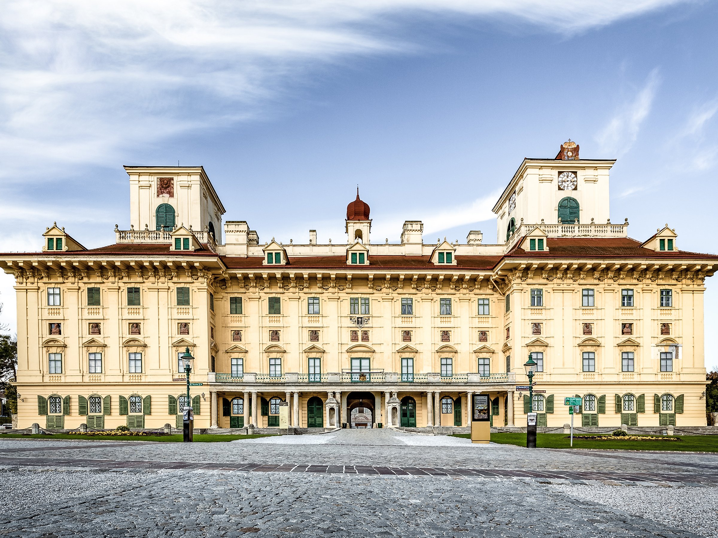 Das Bild zeigt ein prächtiges, kunstvolles Gebäude mit einer kunstvollen Fassade, symmetrischem Design und einem zentralen, gewölbten Eingang. Es verfügt über drei Türme mit Uhren und steht vor einem strahlend blauen Himmel.