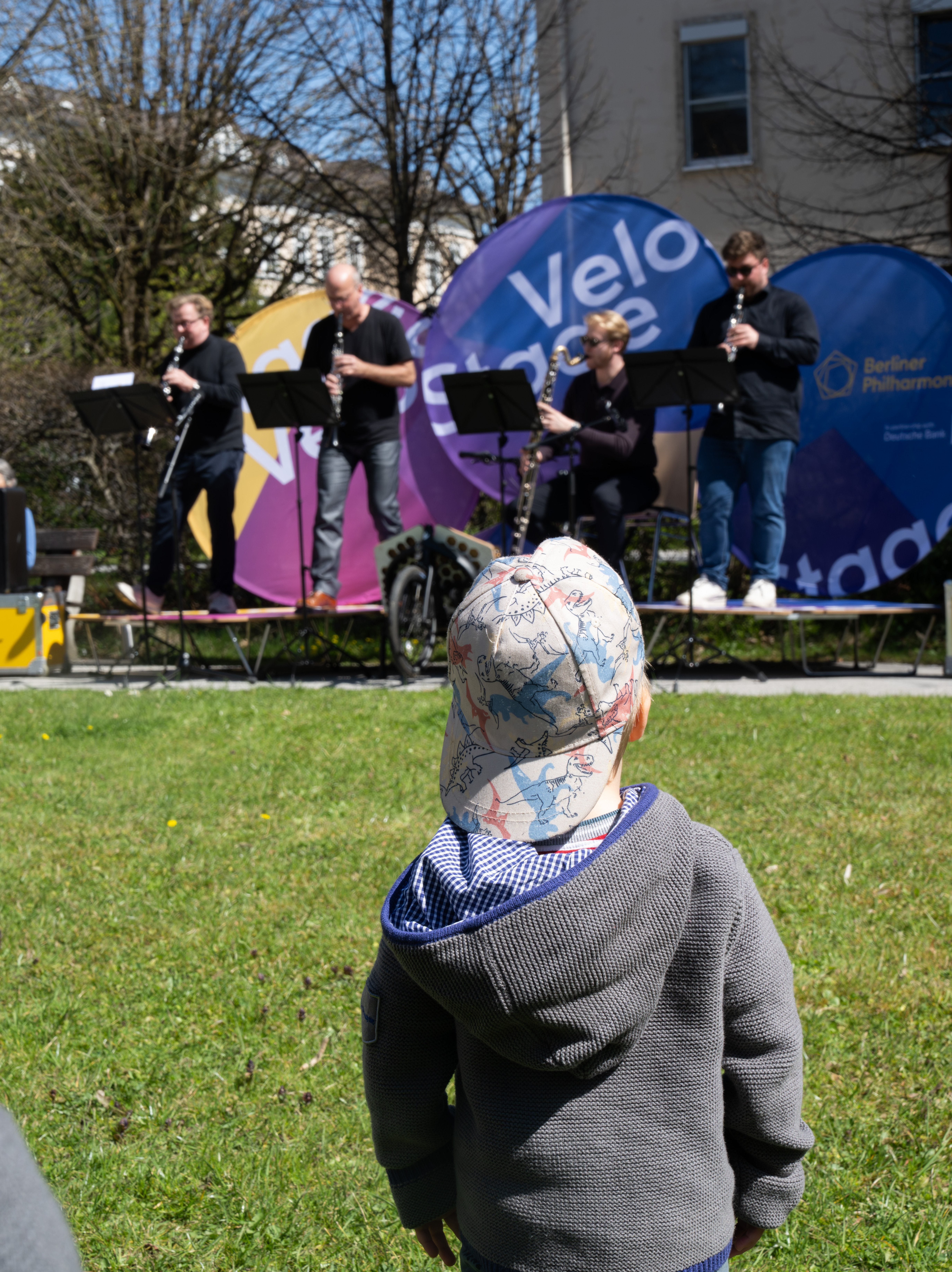 A young child in a hoodie stands on grass, watching a small band of four musicians performing on an outdoor stage with colorful banners in the background on a sunny day.