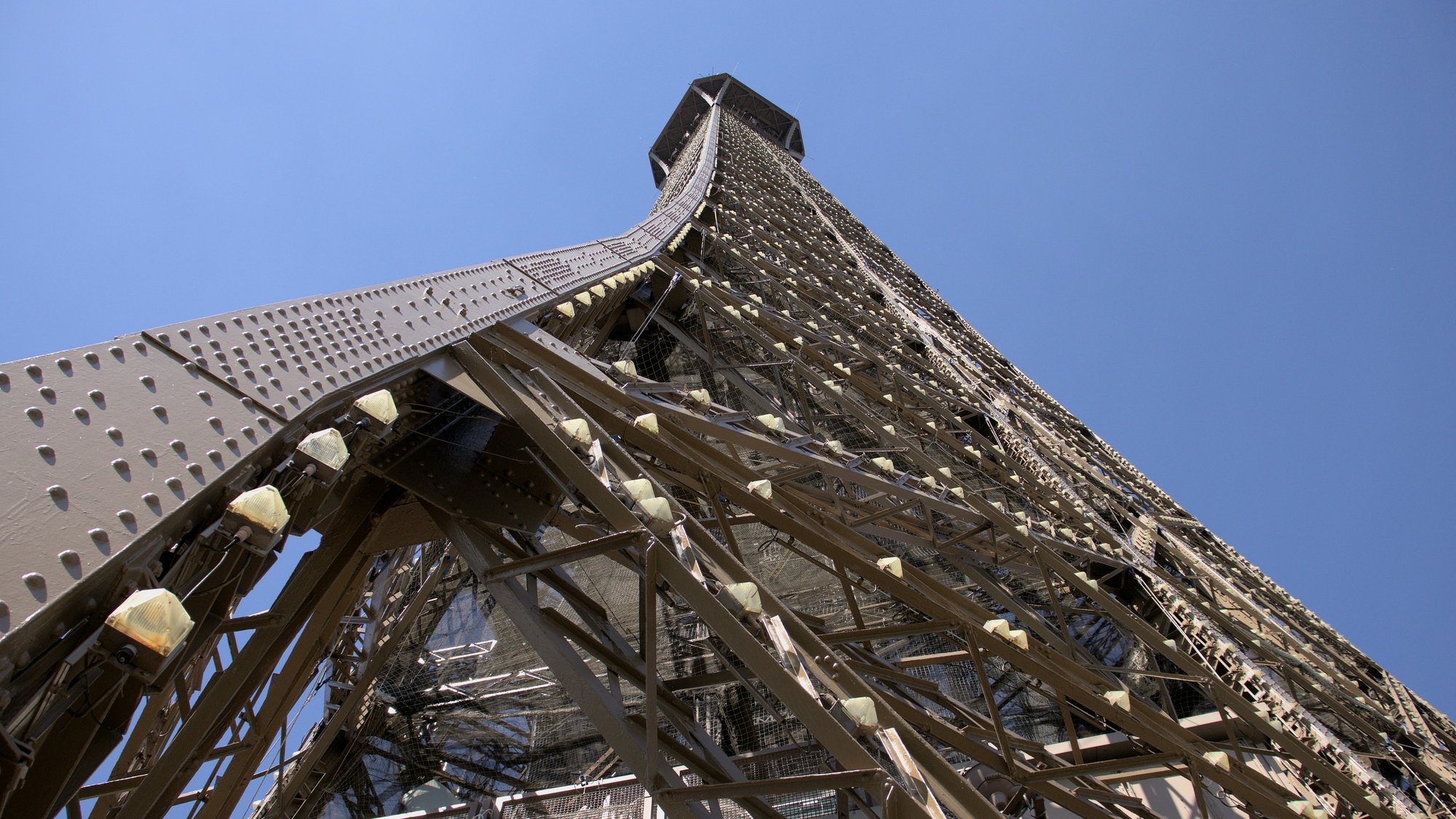 Nahaufnahme des Eiffelturms von unten, mit Blick nach oben entlang seiner Metallstruktur vor einem klaren blauen Himmel. Die komplizierten Eisenarbeiten und Nieten sind im starken Sonnenlicht sichtbar.