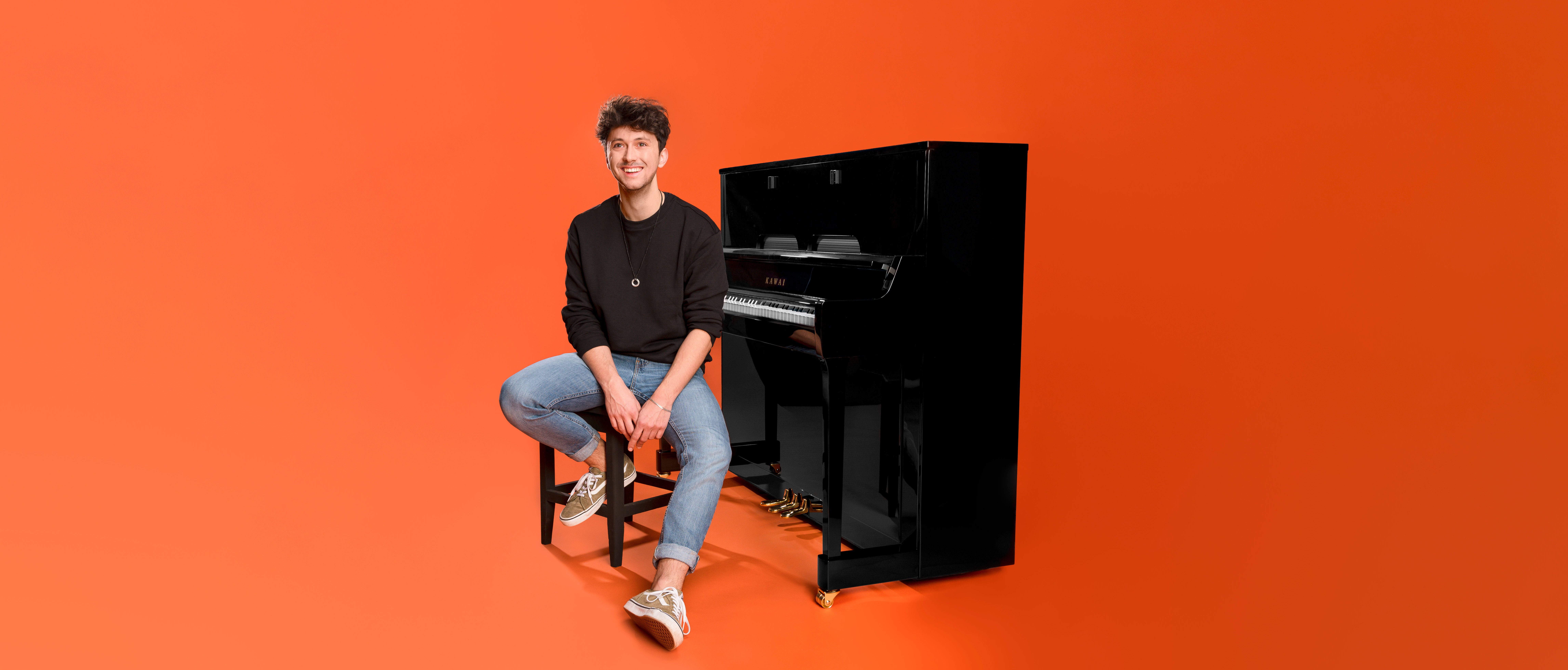 Eine Person in einem schwarzen Pullover und Jeans sitzt auf einem Hocker neben einem schwarzen Klavier vor einem einfarbigen orangefarbenen Hintergrund und lächelt in die Kamera.