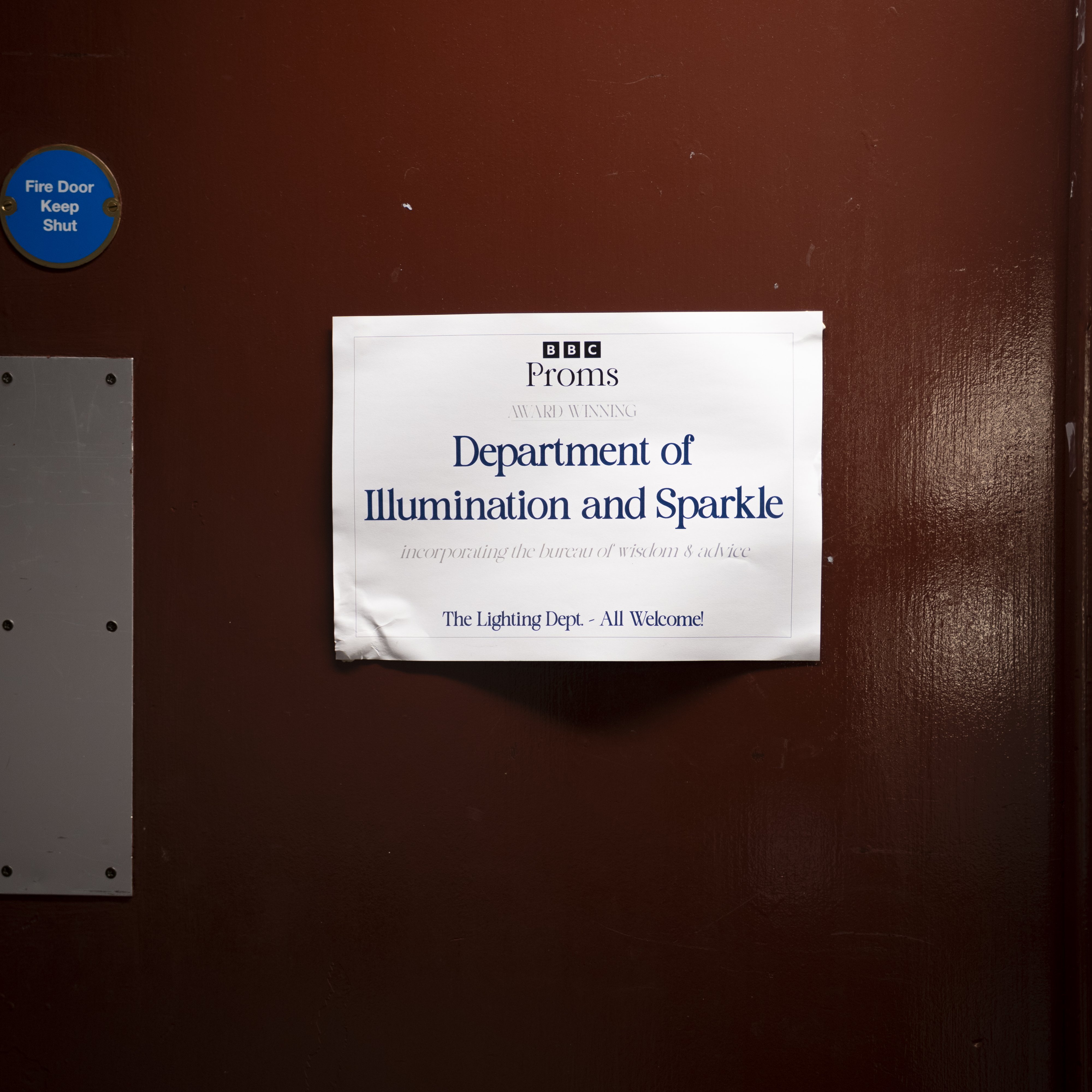 Auf einem weißen Schild an einer braunen Tür steht: BBC Proms – Abteilung für Beleuchtung und Glanz. Die Beleuchtungsabteilung – Alle sind herzlich willkommen! Auf einem blauen, runden Schild steht: „Feuertür – Bitte geschlossen halten».