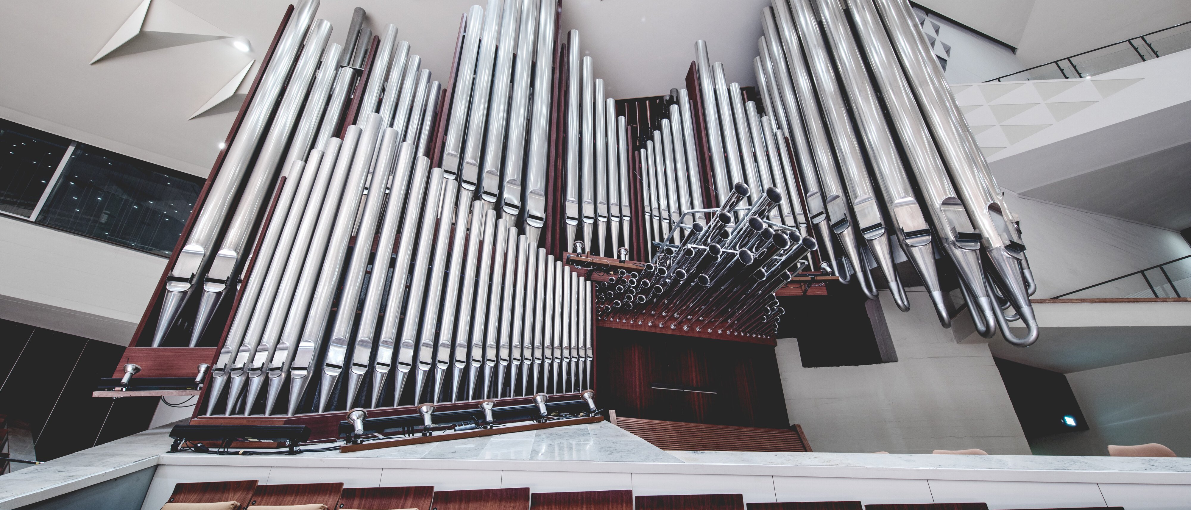 A large pipe organ with numerous shiny metal pipes is mounted at the front of a modern concert hall, with empty wooden seats and a white ceiling featuring geometric designs.
