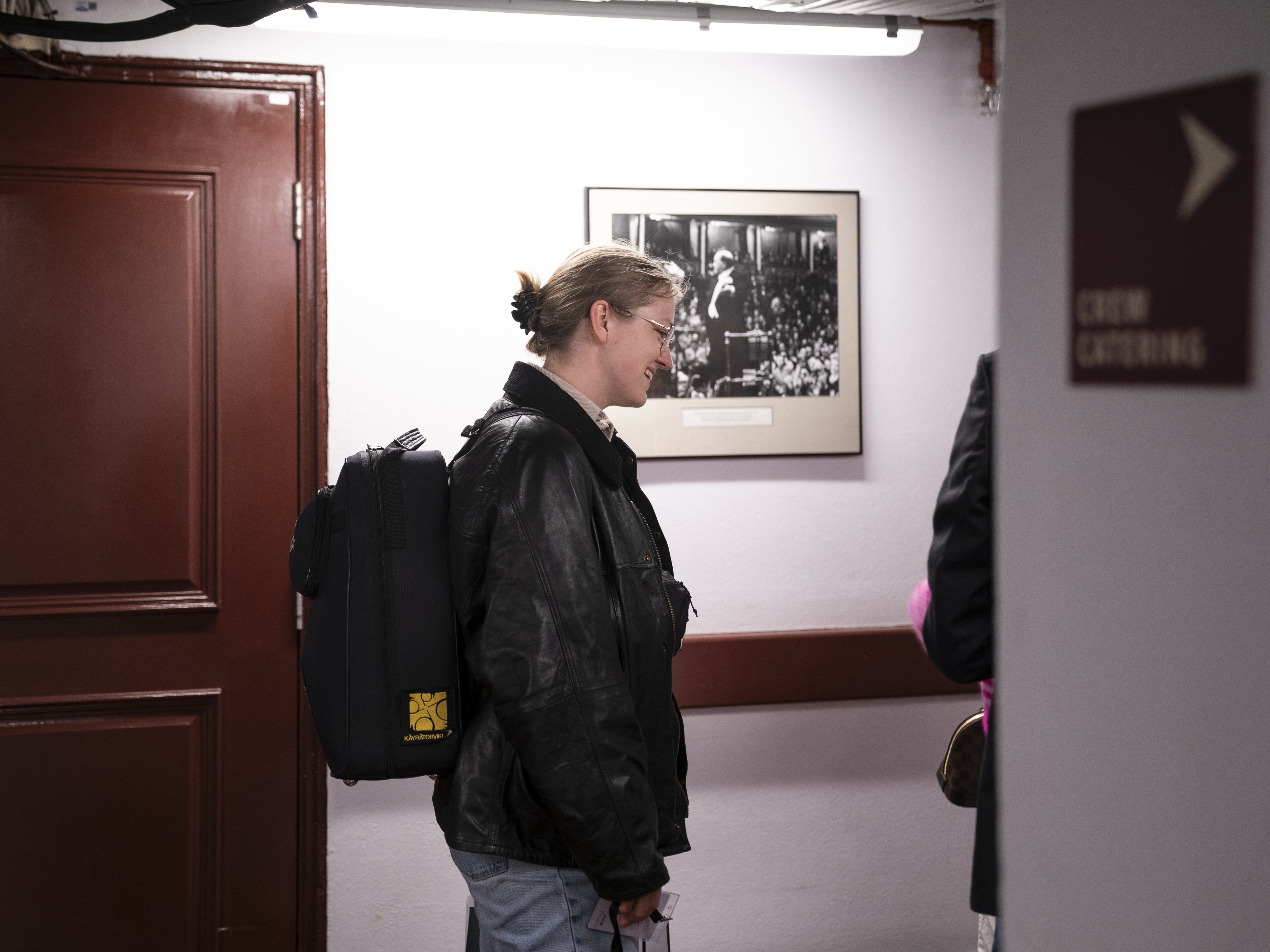 Eine Person mit Brille und schwarzer Lederjacke steht mit einem schwarzen Rucksack in der Hand neben einer roten Tür. An der Wand hinter ihr hängt ein Schwarz-Weiß-Foto.