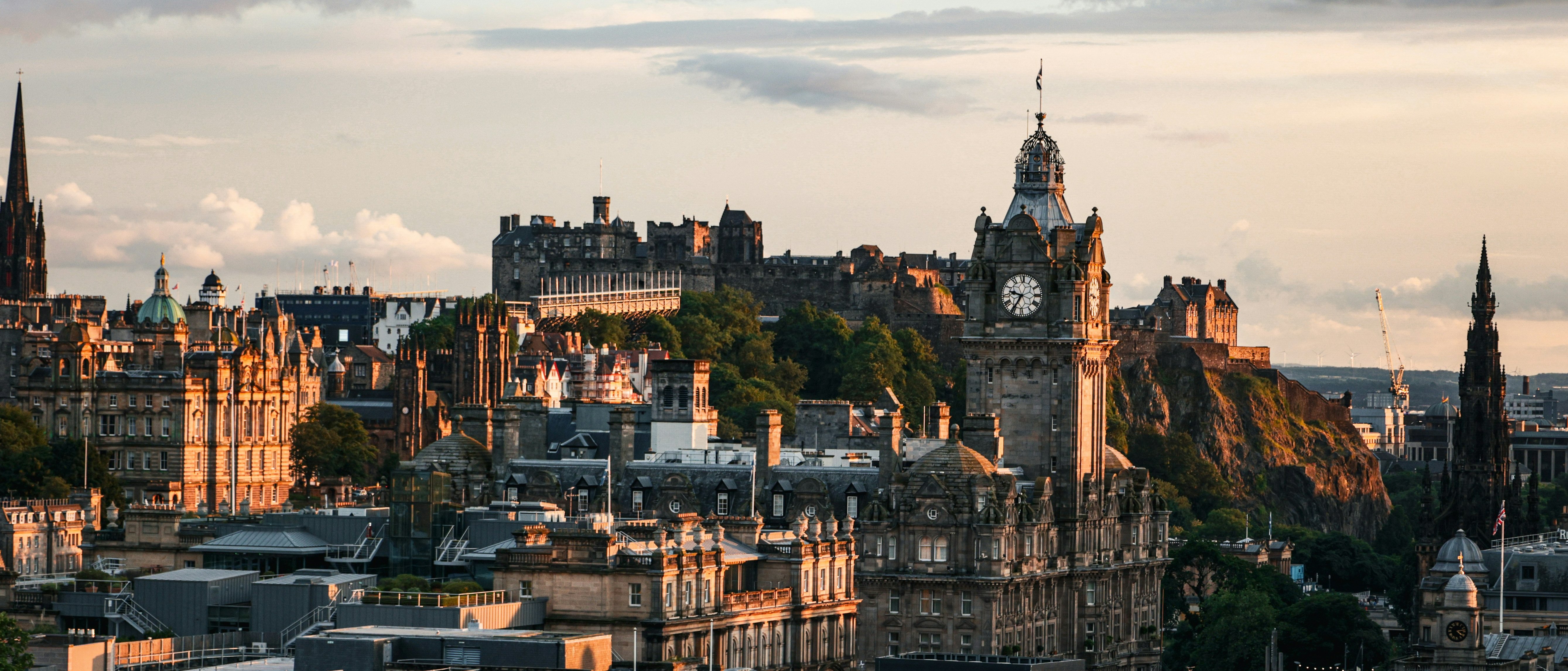 Edinburgh cityscape at sunset, featuring the historic Edinburgh Castle on a hill, the Balmoral Hotel clock tower, and various old stone buildings under a partly cloudy sky.