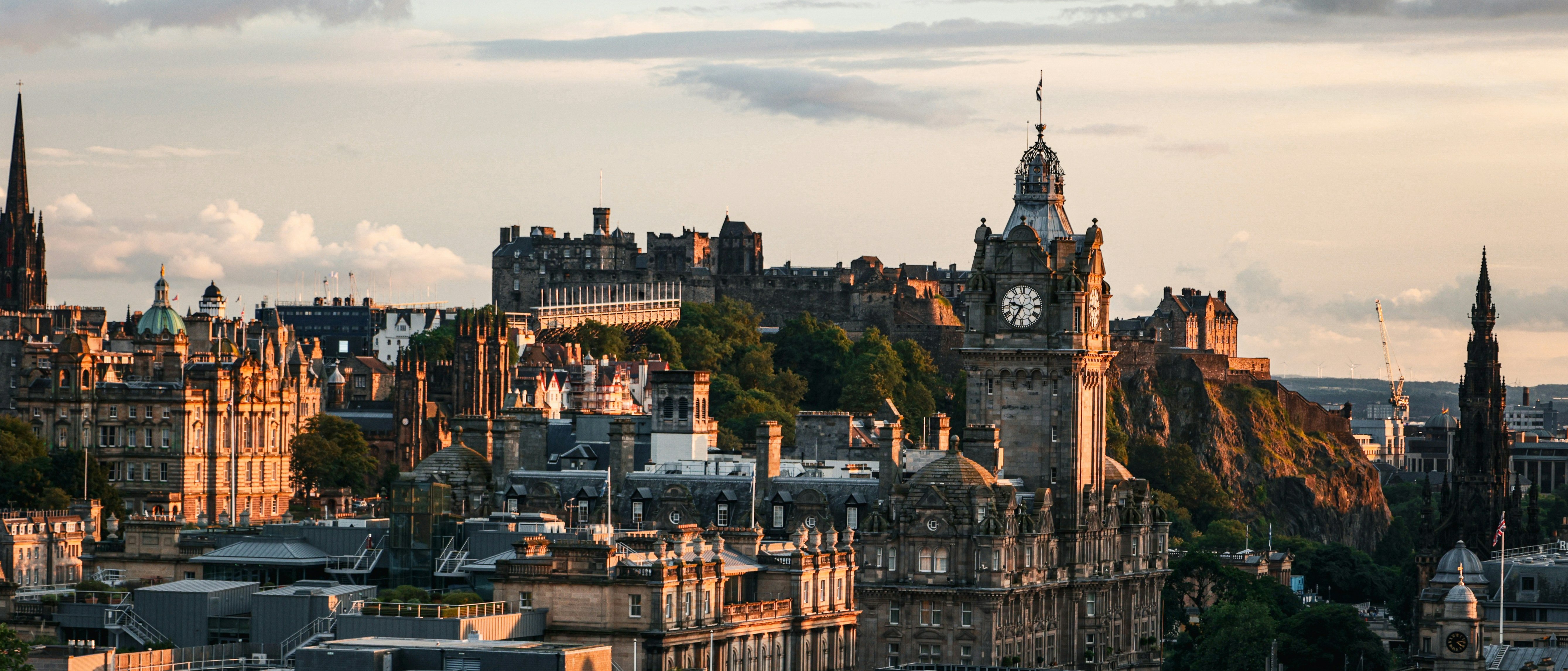 Stadtansicht von Edinburgh bei Sonnenuntergang mit dem Uhrturm des Balmoral Hotels, historischen Gebäuden und dem Edinburgh Castle auf einem Hügel im Hintergrund unter bewölktem Himmel.