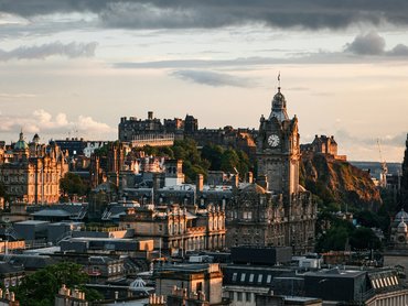 Stadtansicht von Edinburgh bei Sonnenuntergang mit dem Uhrturm des Balmoral Hotels, historischen Gebäuden und dem Edinburgh Castle auf einem Hügel im Hintergrund unter bewölktem Himmel.
