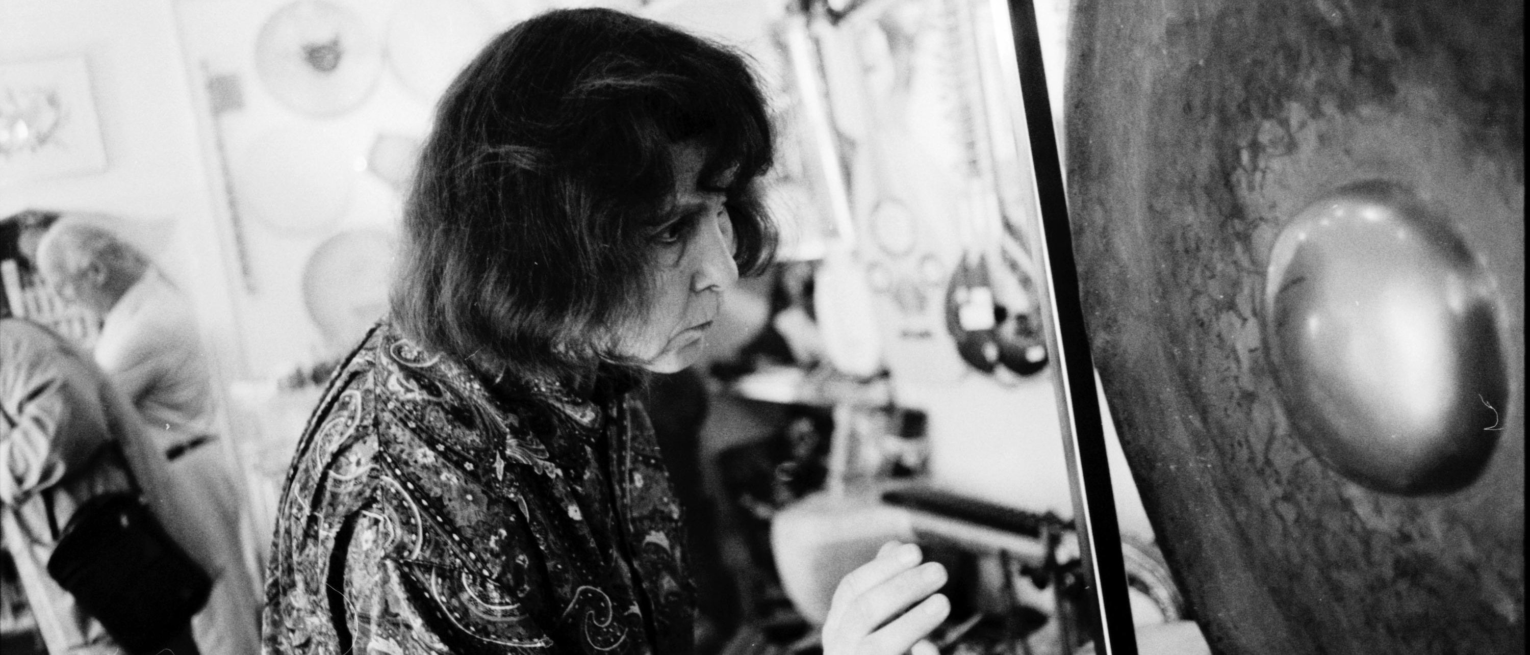 A person with shoulder-length hair and a patterned shirt is striking a large gong in a room filled with instruments and hanging decorations. The scene is captured in black and white.