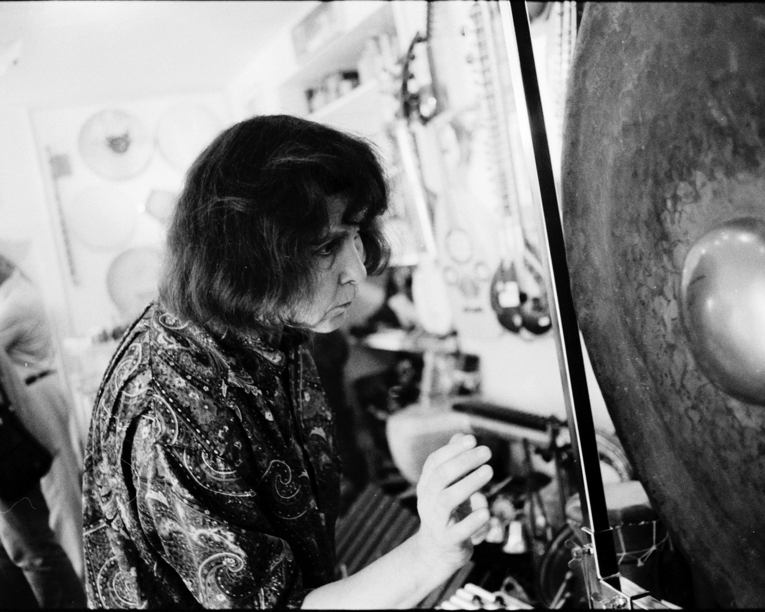 Eine Person mit mittellangem Haar und einem gemusterten Hemd spielt sanft einen großen Gong in einem Raum, der mit Musikinstrumenten und verschiedenen hängenden Gegenständen gefüllt ist. Das Foto ist in schwarz-weiß gehalten.