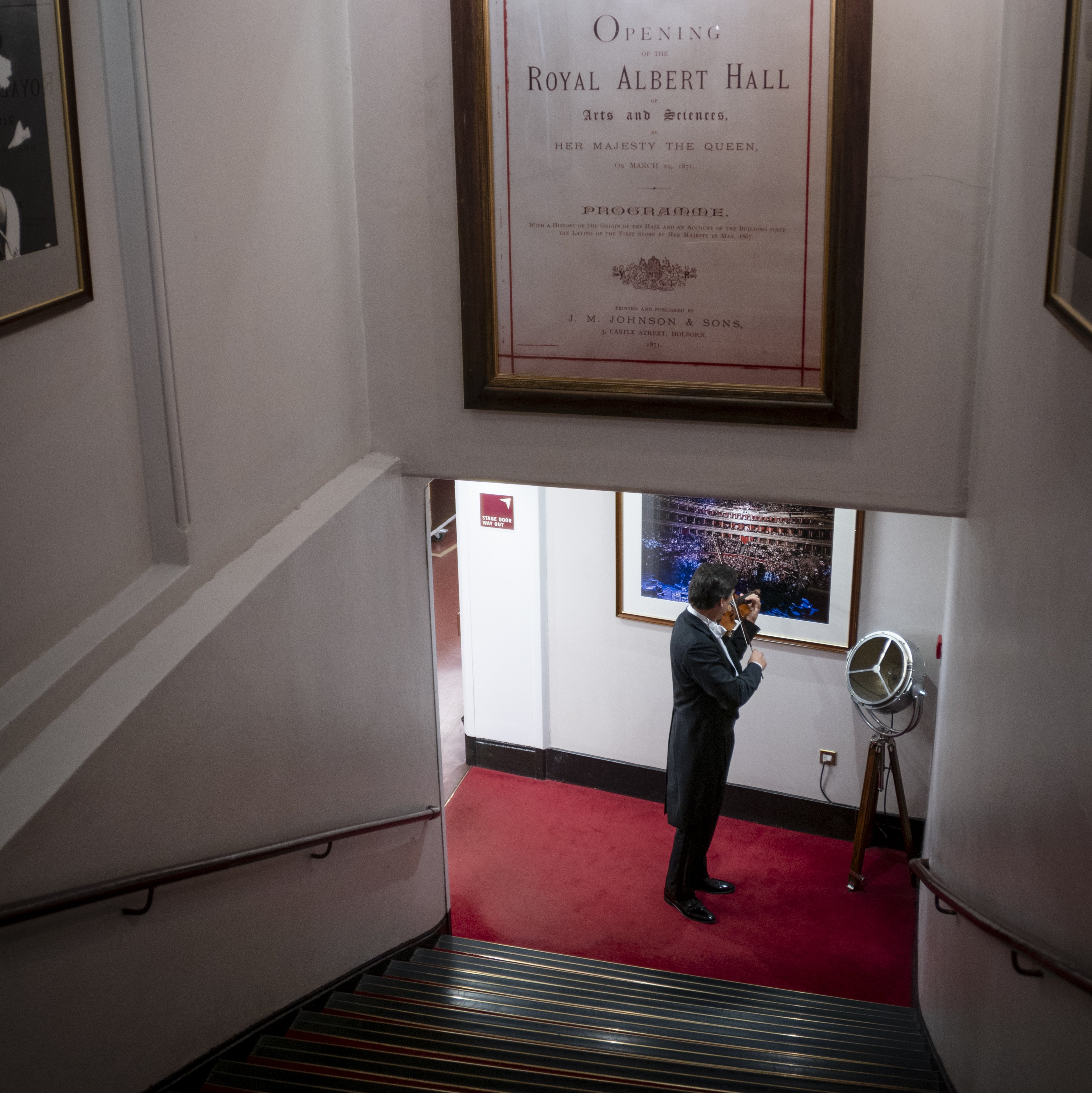 Ein Mann im Anzug steht auf einem mit rotem Teppich ausgelegten Treppenabsatz und telefoniert unter einem großen, gerahmten Plakat zur Eröffnung der Royal Albert Hall. Treppen und Kunstwerke umrahmen die Szene.