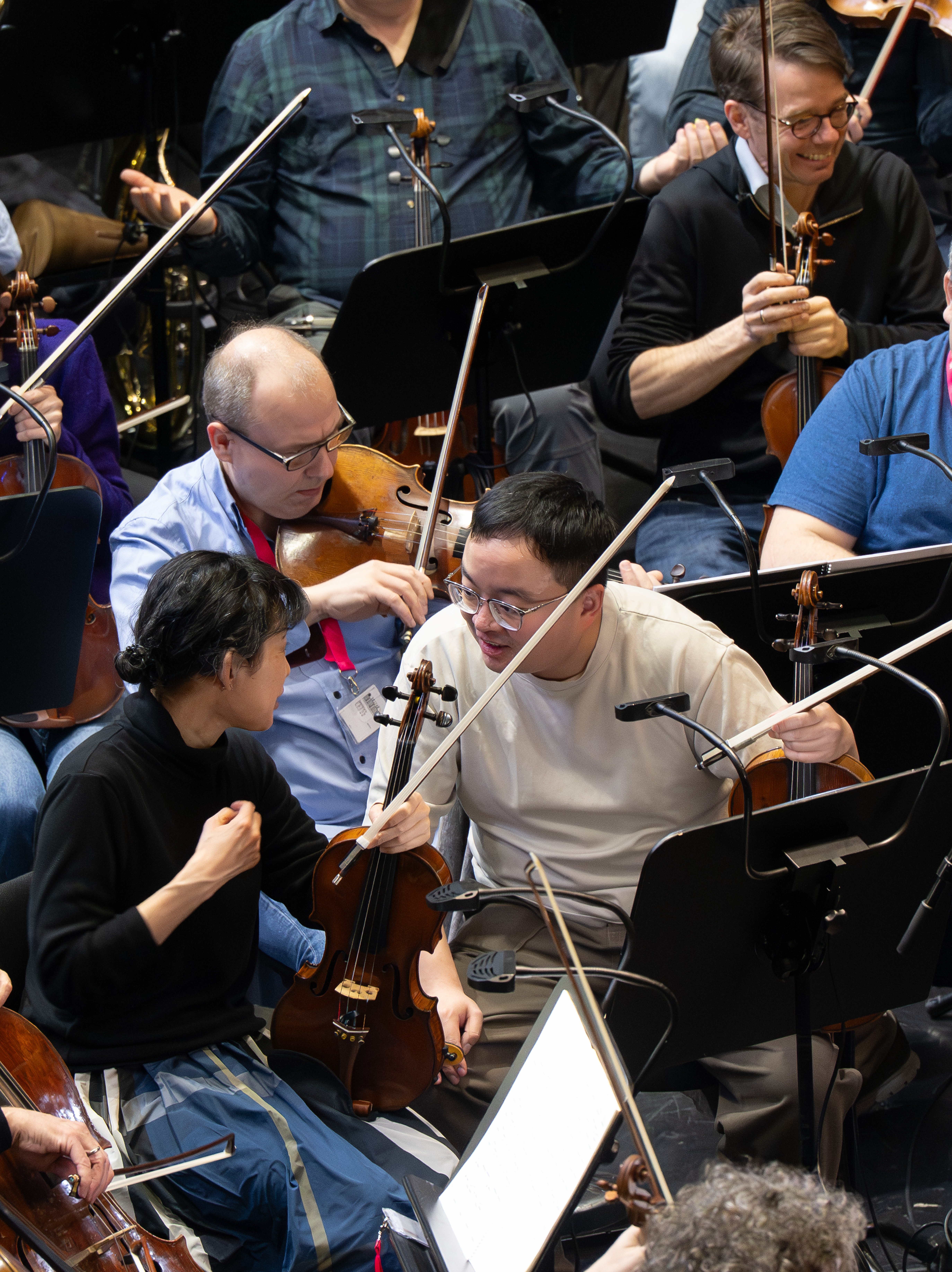 Die Musiker eines Orchesters, die dicht neben ihren Streichinstrumenten sitzen, unterhalten sich während einer Probe. Die Notenblätter sind auf Ständern vor ihnen zu sehen.