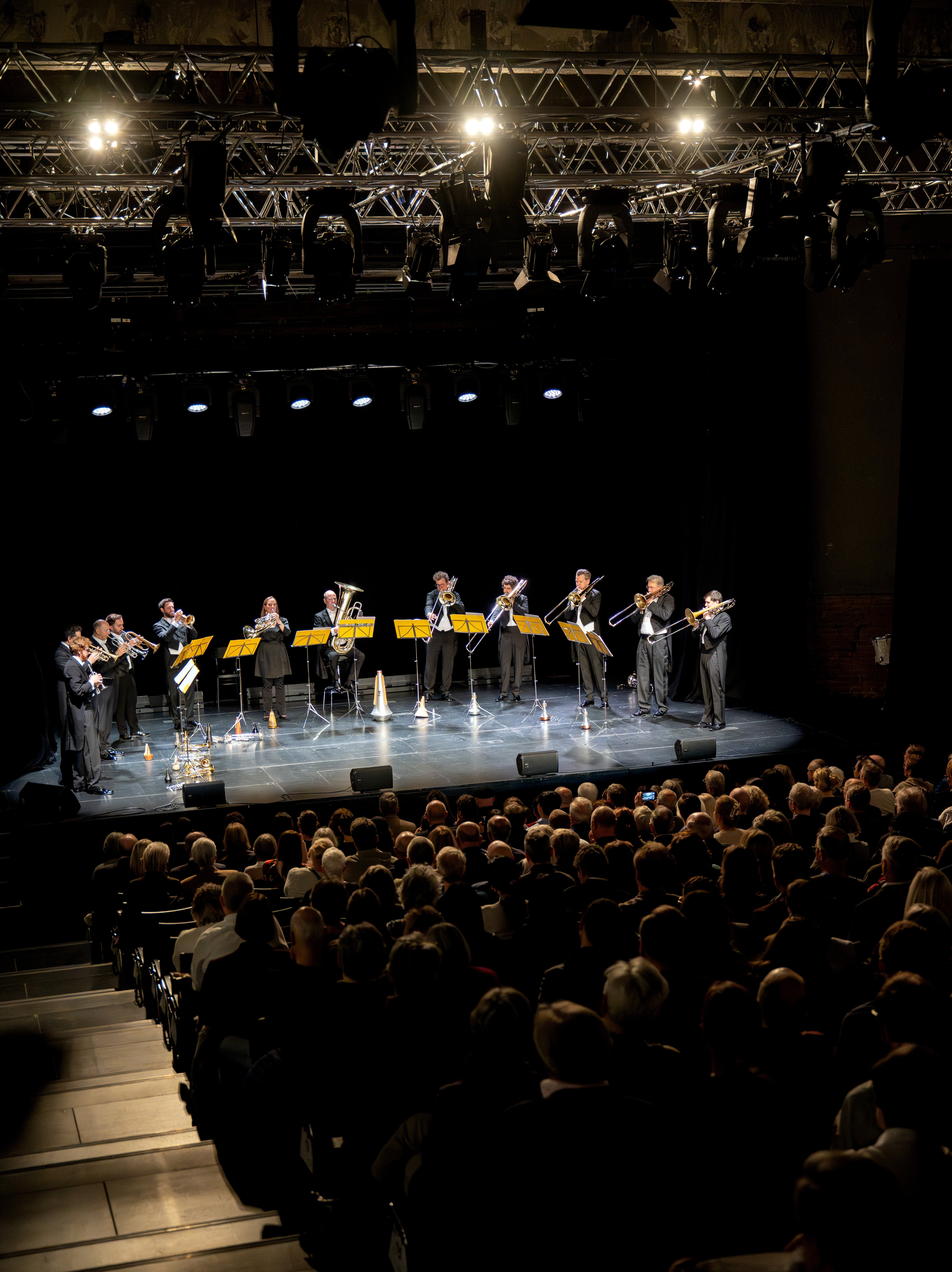 Ein Blechbläserensemble spielt auf einer Bühne unter hellen Lichtern, gegenüber einem sitzenden Publikum in einem abgedunkelten Theater. Die Musiker stehen in einer Reihe mit Notenständern und Instrumenten, gekleidet in formelle schwarze Kleidung.