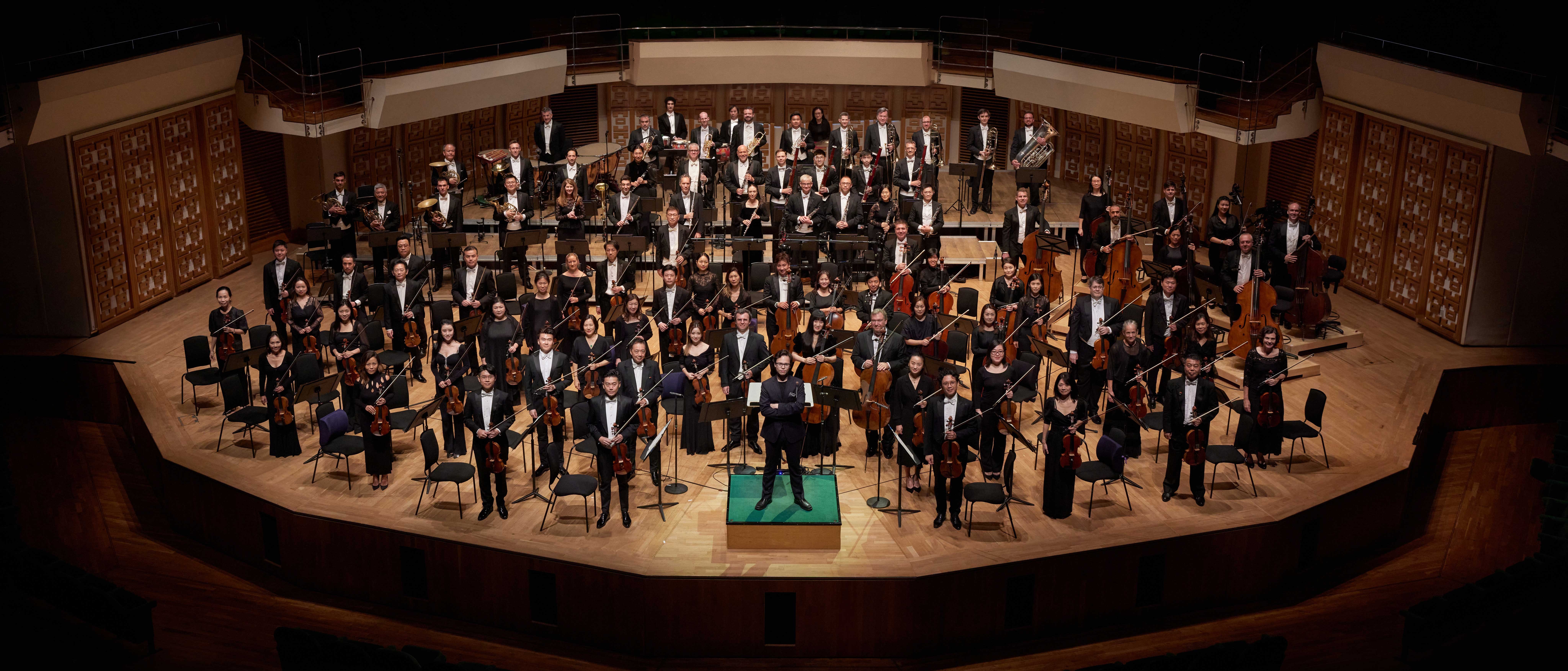 Auf der Bühne eines Konzertsaals sitzt ein vollbesetztes Orchester; die Musiker halten verschiedene Instrumente in den Händen, und in der Mitte des Podiums steht ein Dirigent. Der Saal ist leer und bis auf die Bühne nur schwach beleuchtet.
