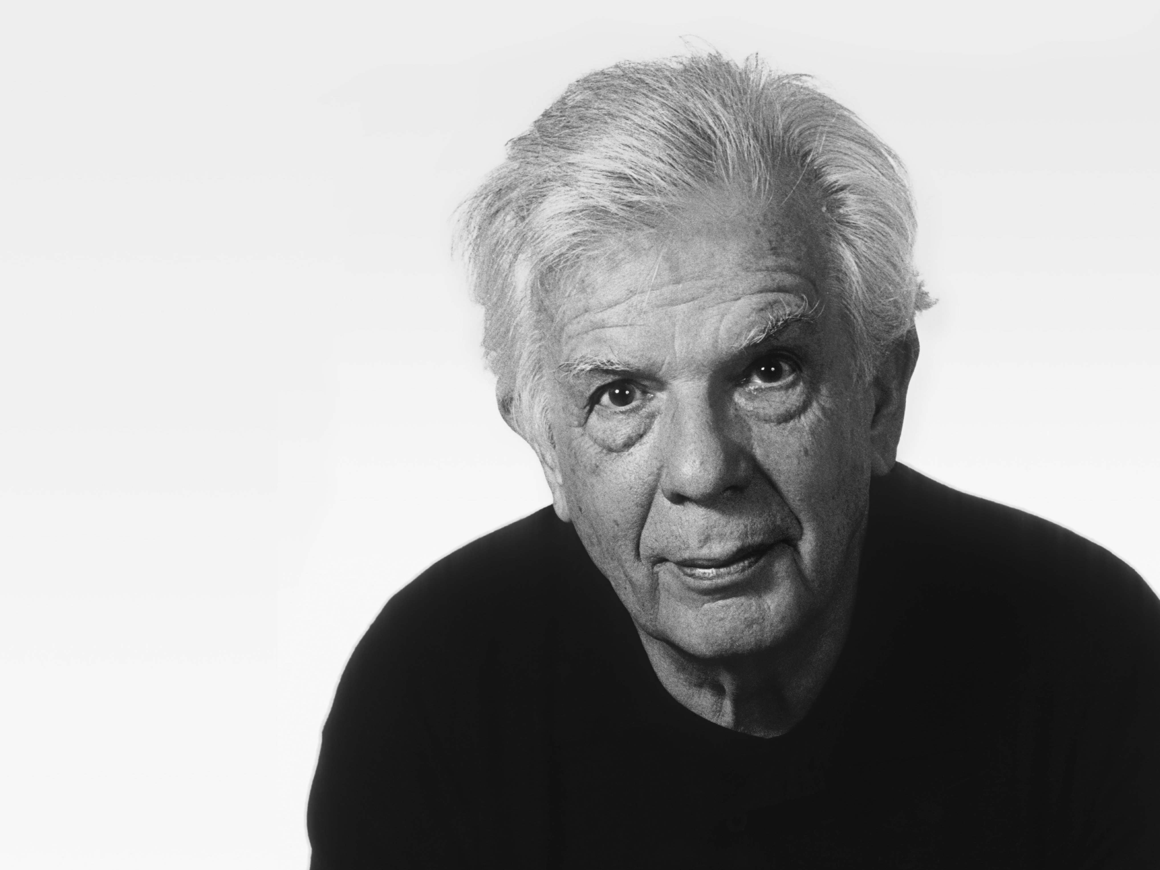 Black and white portrait of an older man with white hair, wearing a dark shirt, looking directly at the camera with a neutral expression against a plain light background.