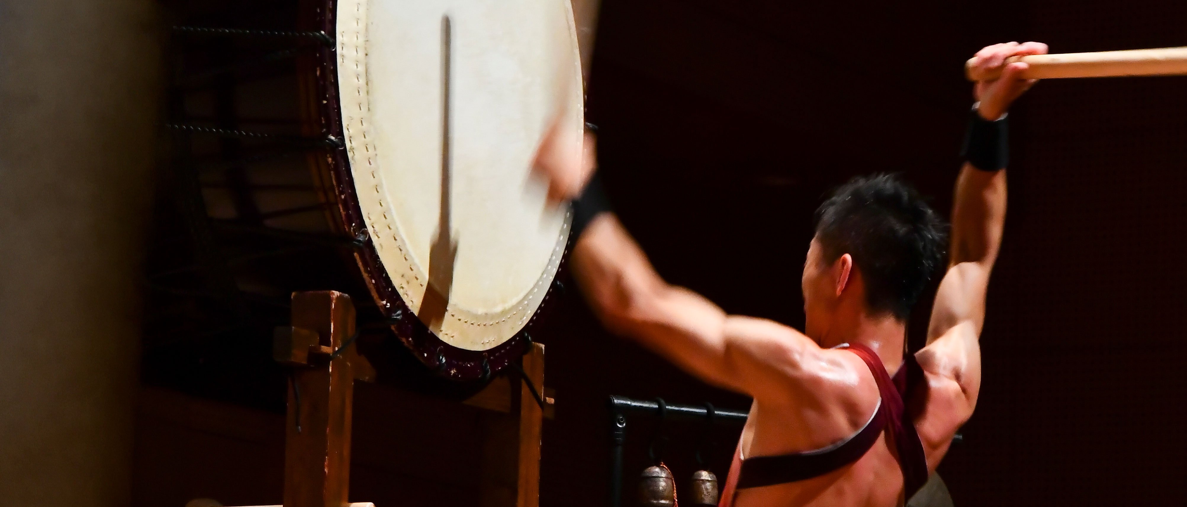 Ein muskulöser Schlagzeuger mit nacktem Rücken spielt in einem schummrig beleuchteten Veranstaltungsraum auf einer großen traditionellen japanischen Taiko-Trommel und hält die Trommelstöcke dabei hoch.