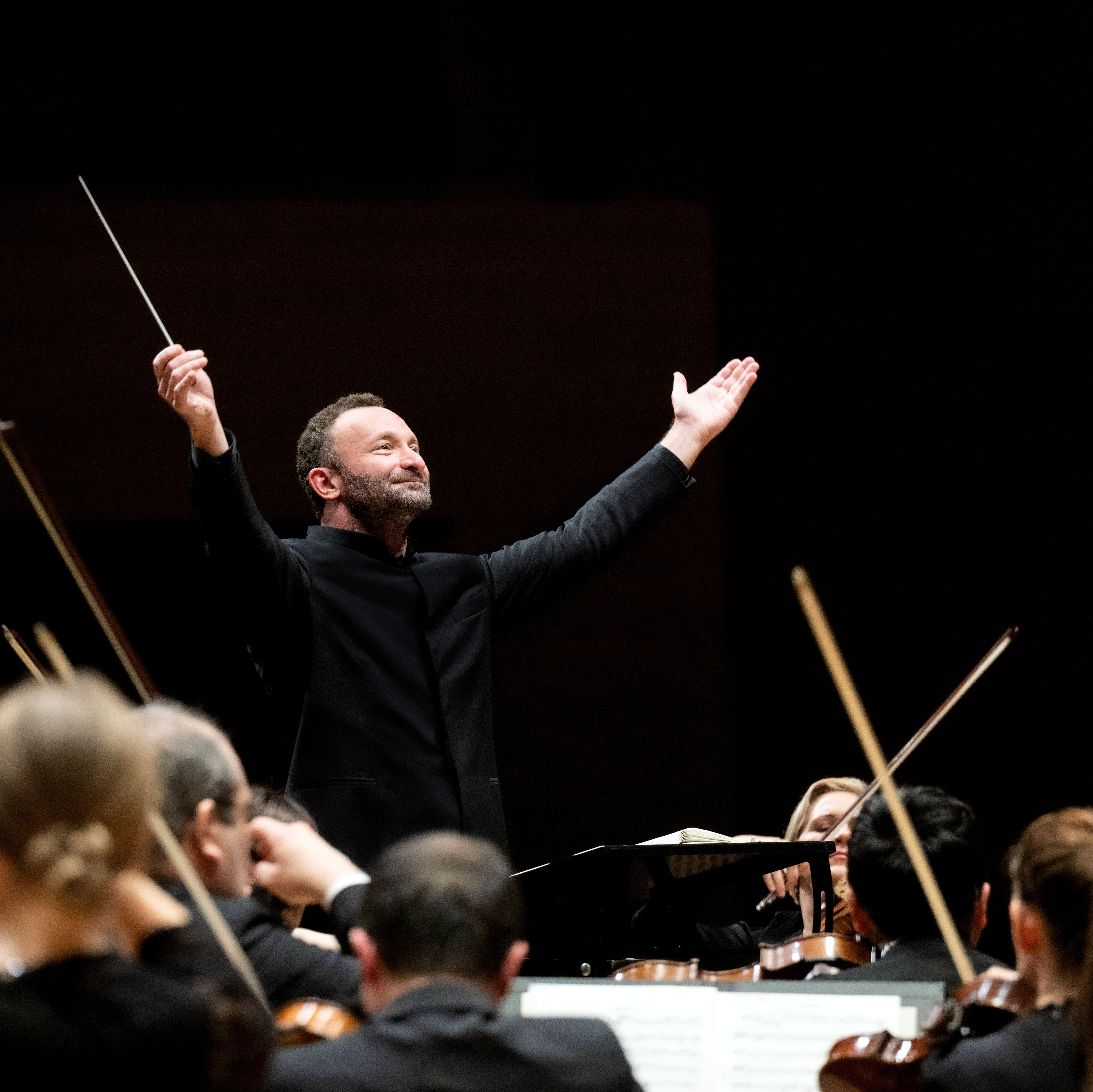 Kirill Petrenko steht vor dem Orchester und hebt beide Arme zu einer großen Geste nach oben. Er trägt einen schwarzen Anzug. Im Vordergrund sind Musiker von hinten zu sehen.