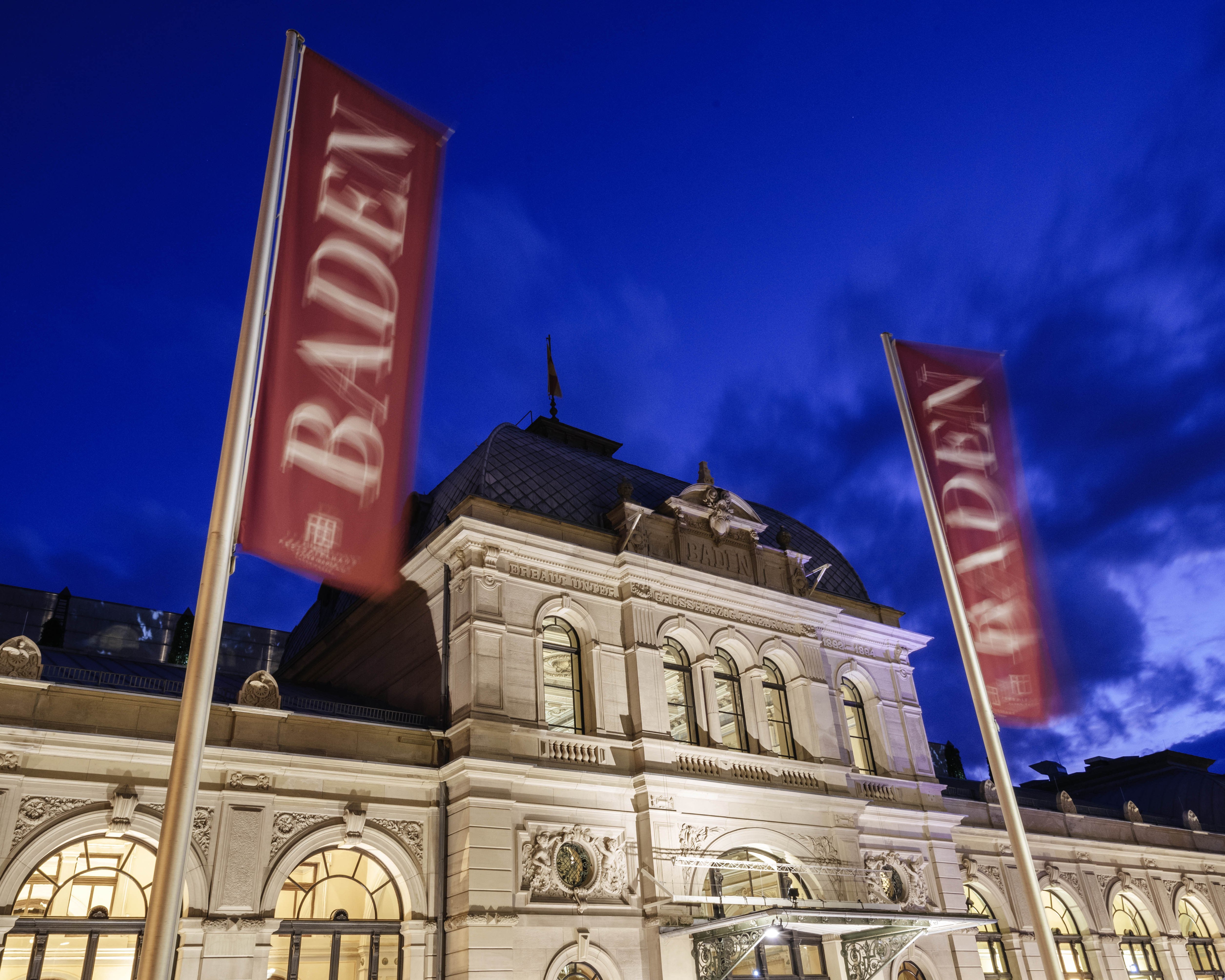 Historic building illuminated at night with two red flags reading “BADEN” in front, set against a deep blue sky with scattered clouds. Warm light shines through arched windows.