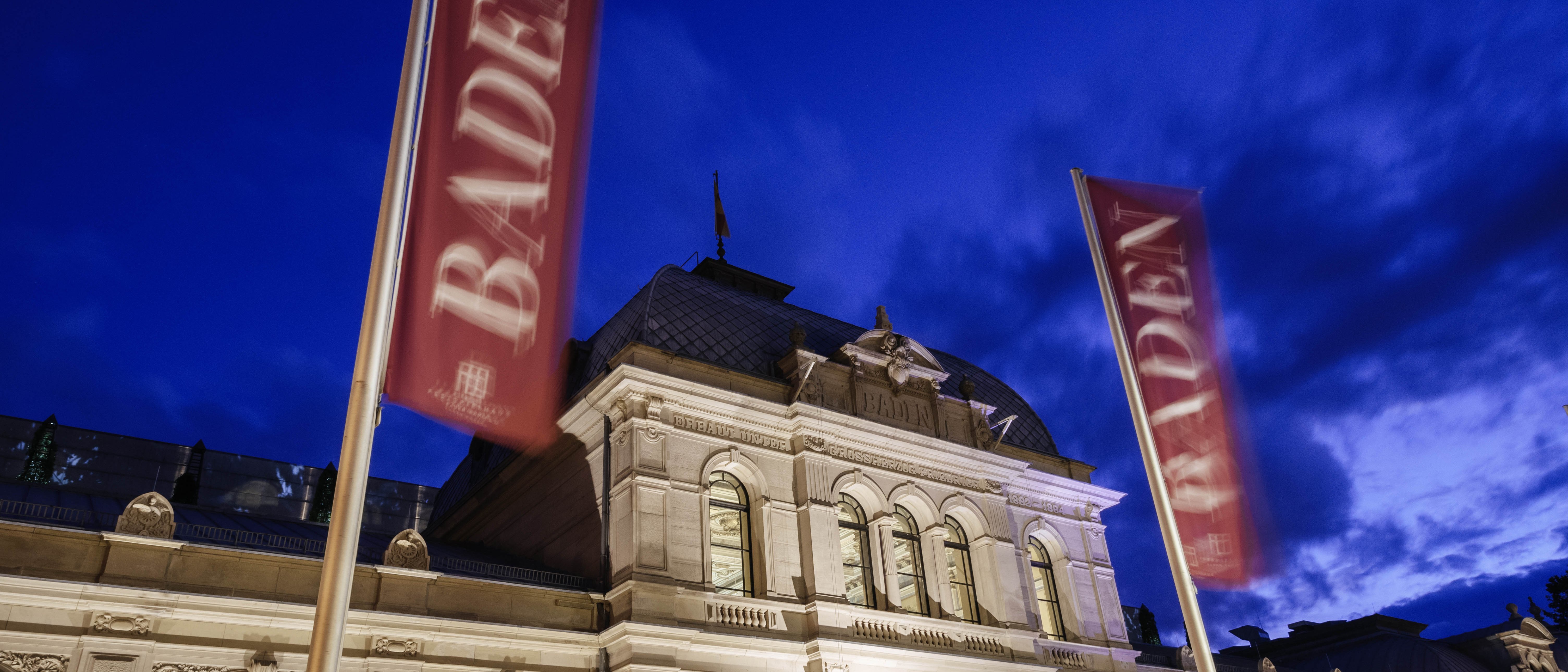 Elegantes historisches Gebäude, nachts beleuchtet, mit zwei roten Fahnen mit der Aufschrift BADEN davor, vor einem tiefblauen Himmel mit vereinzelten Wolken.
