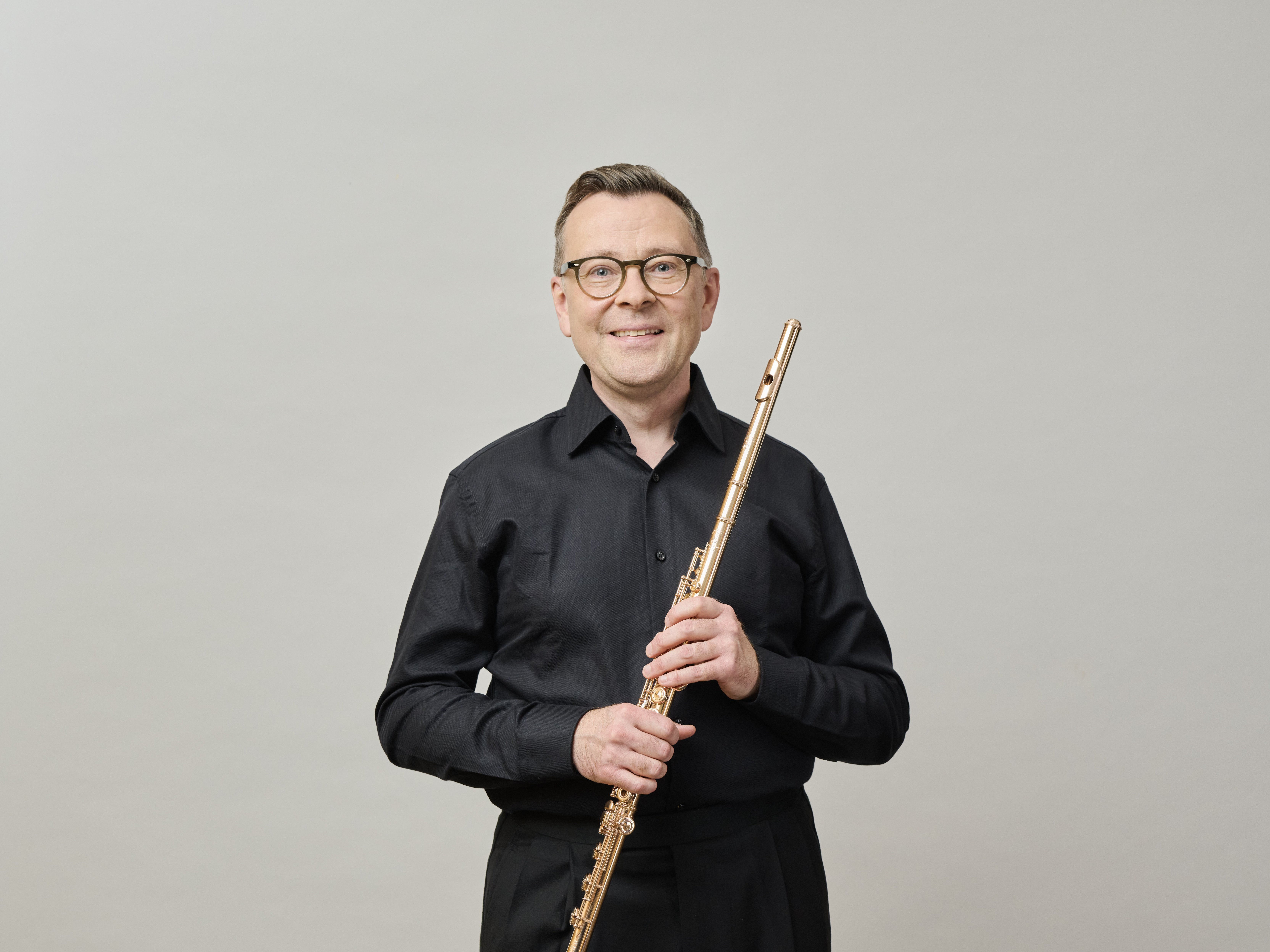 A man wearing glasses and a black shirt stands in front of a plain light background, smiling while holding a flute vertically with both hands.