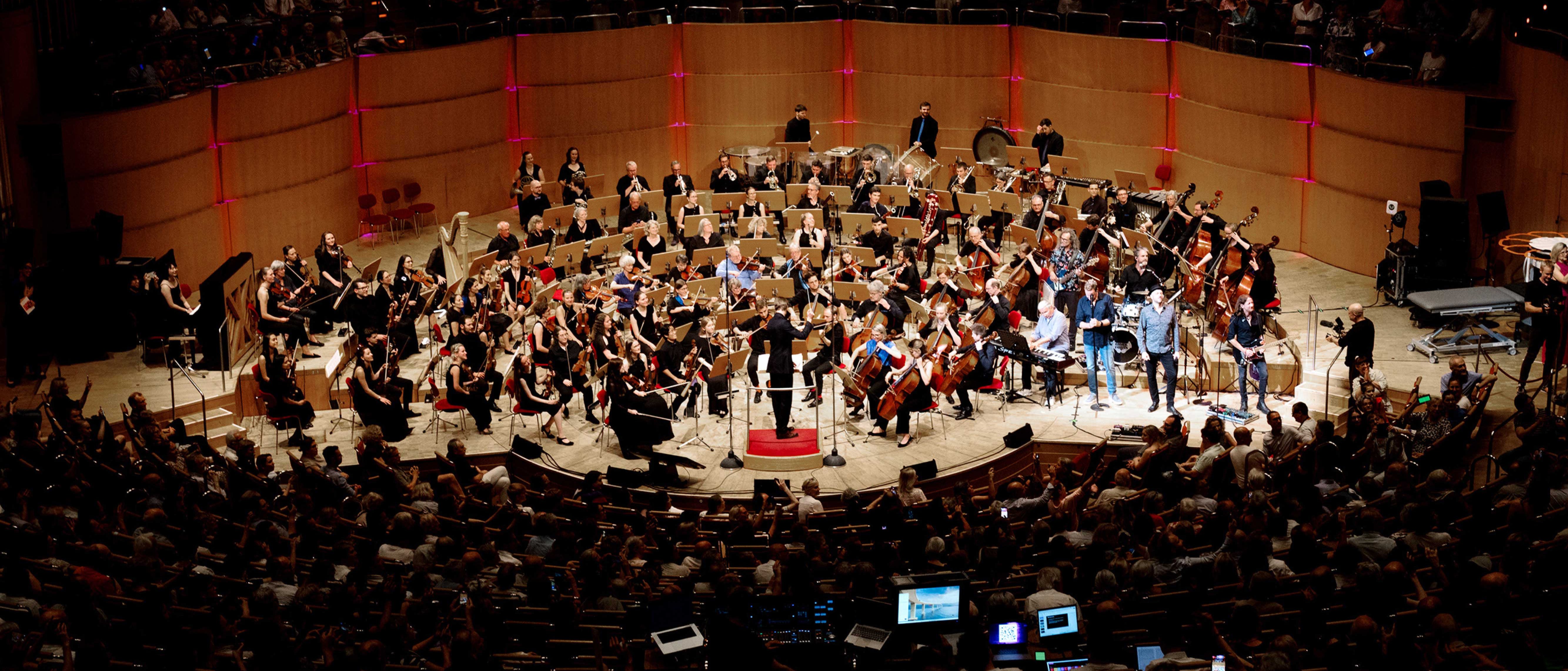 Auf der Bühne eines Konzertsaals spielt ein großes Orchester; die Musiker spielen verschiedene Instrumente, und das Publikum sitzt vor ihnen und um sie herum. Die Bühne ist hell beleuchtet; im Saal ist es größtenteils dunkel.