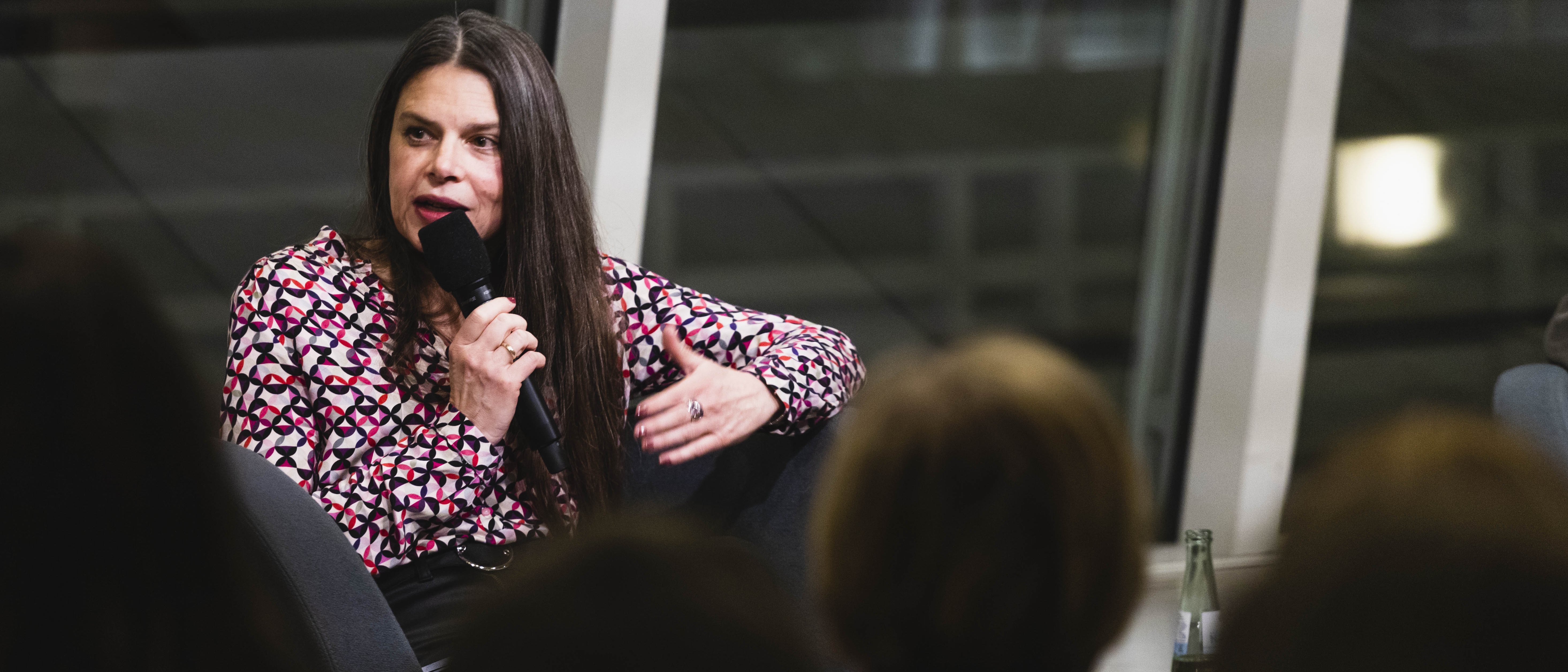 Eine Frau mit langen Haaren und in einer bunten Bluse sitzt in einem Sessel und hält ein Mirkofon in der Hand, in das sie spricht. Vor ihr sitzt Publikum.