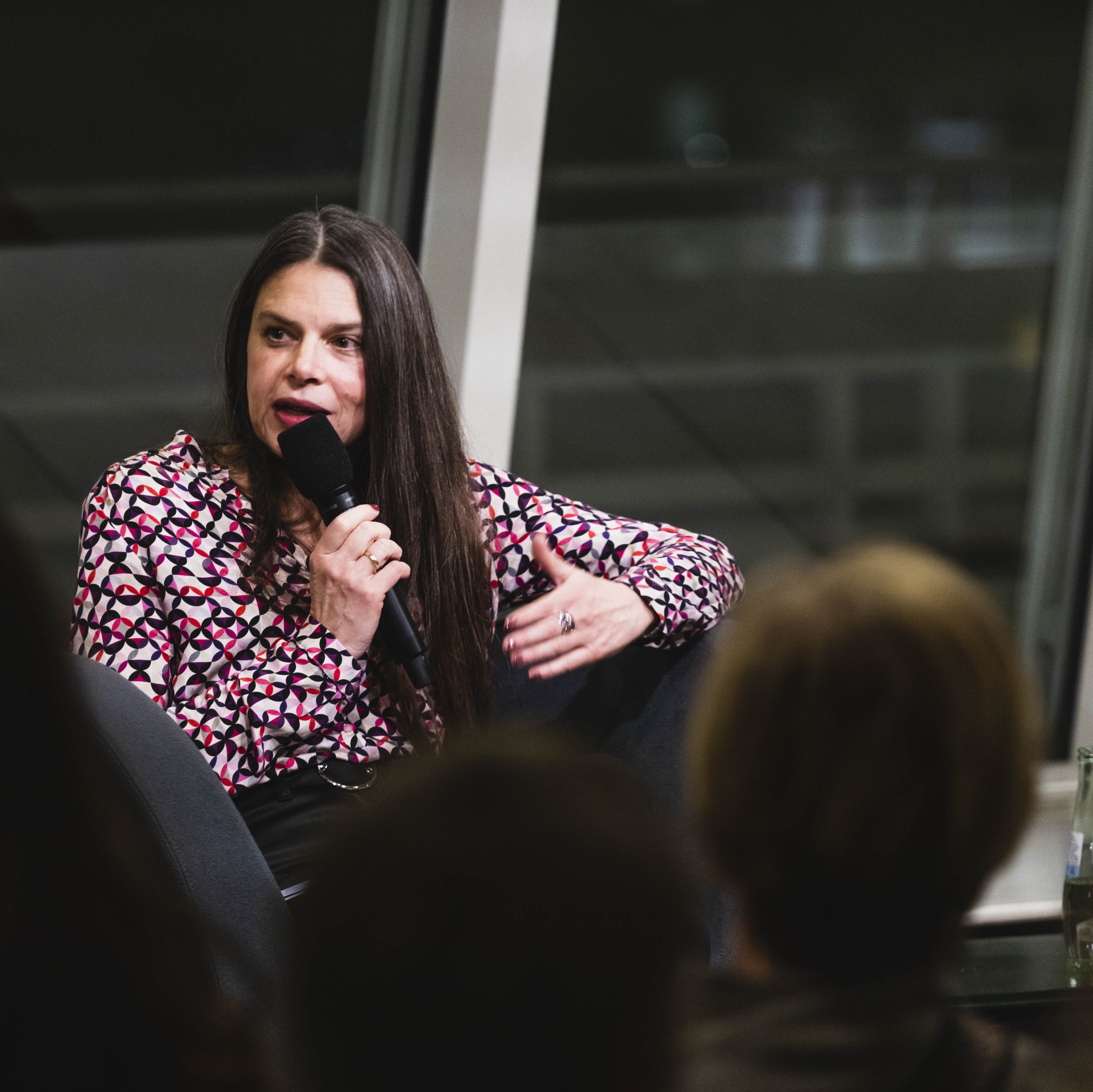 Eine Frau mit langen Haaren und in einer bunten Bluse sitzt in einem Sessel und hält ein Mirkofon in der Hand, in das sie spricht. Vor ihr sitzt Publikum.