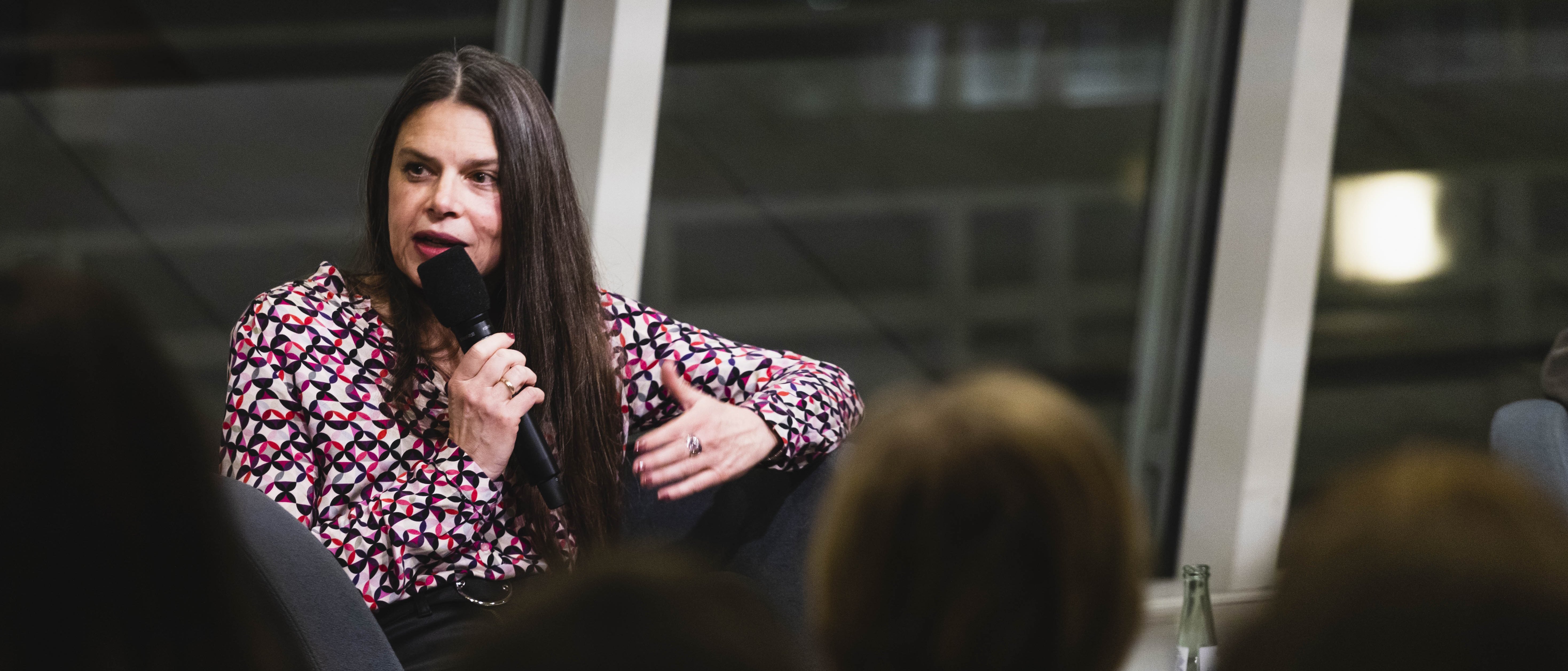 A woman with long hair and wearing a colourful blouse sits in an armchair and holds a microphone in her hand into which she speaks. An audience sits in front of her.