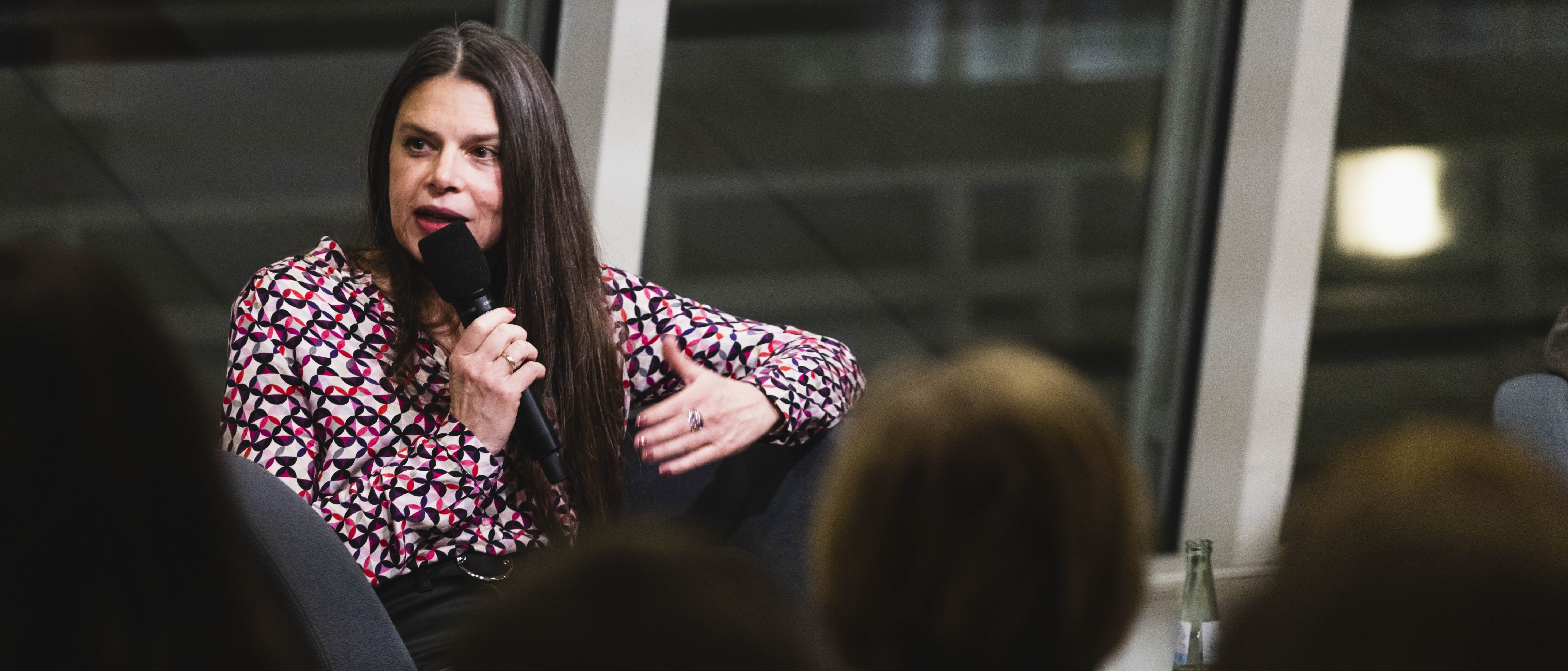 Eine Frau mit langen Haaren und in einer bunten Bluse sitzt in einem Sessel und hält ein Mirkofon in der Hand, in das sie spricht. Vor ihr sitzt Publikum.