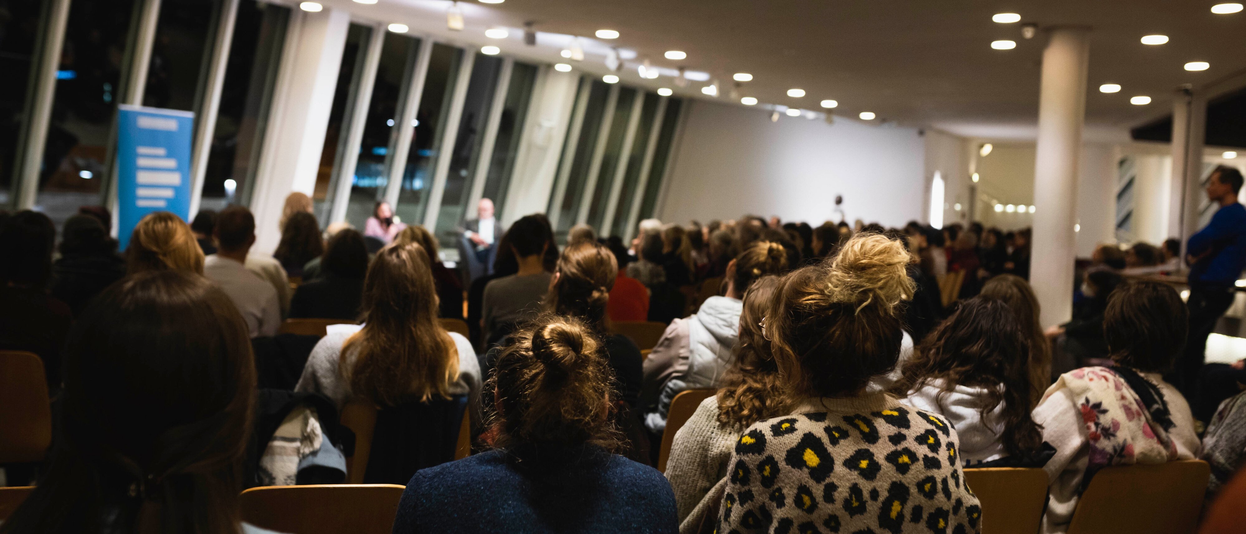 Ein großes Publikum sitzt drinnen vor einer Bühne, auf der die Redner eine Diskussion führen. Der Raum hat hohe Fenster, eine moderne Beleuchtung und Menschen verschiedenen Alters, die aufmerksam zuhören.