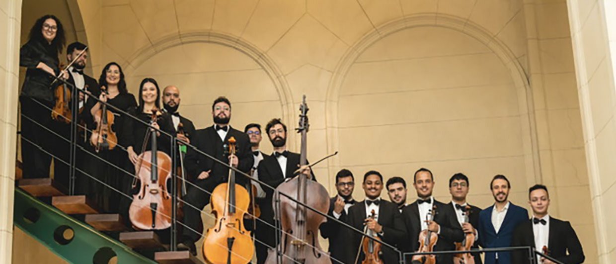 Eine Gruppe von Orchestermusikern in festlicher Kleidung steht auf einer Treppe in einem prächtigen Gebäude, hält verschiedene Streichinstrumente wie Geigen, Celli und einen Kontrabass in den Händen und lächelt in die Kamera.