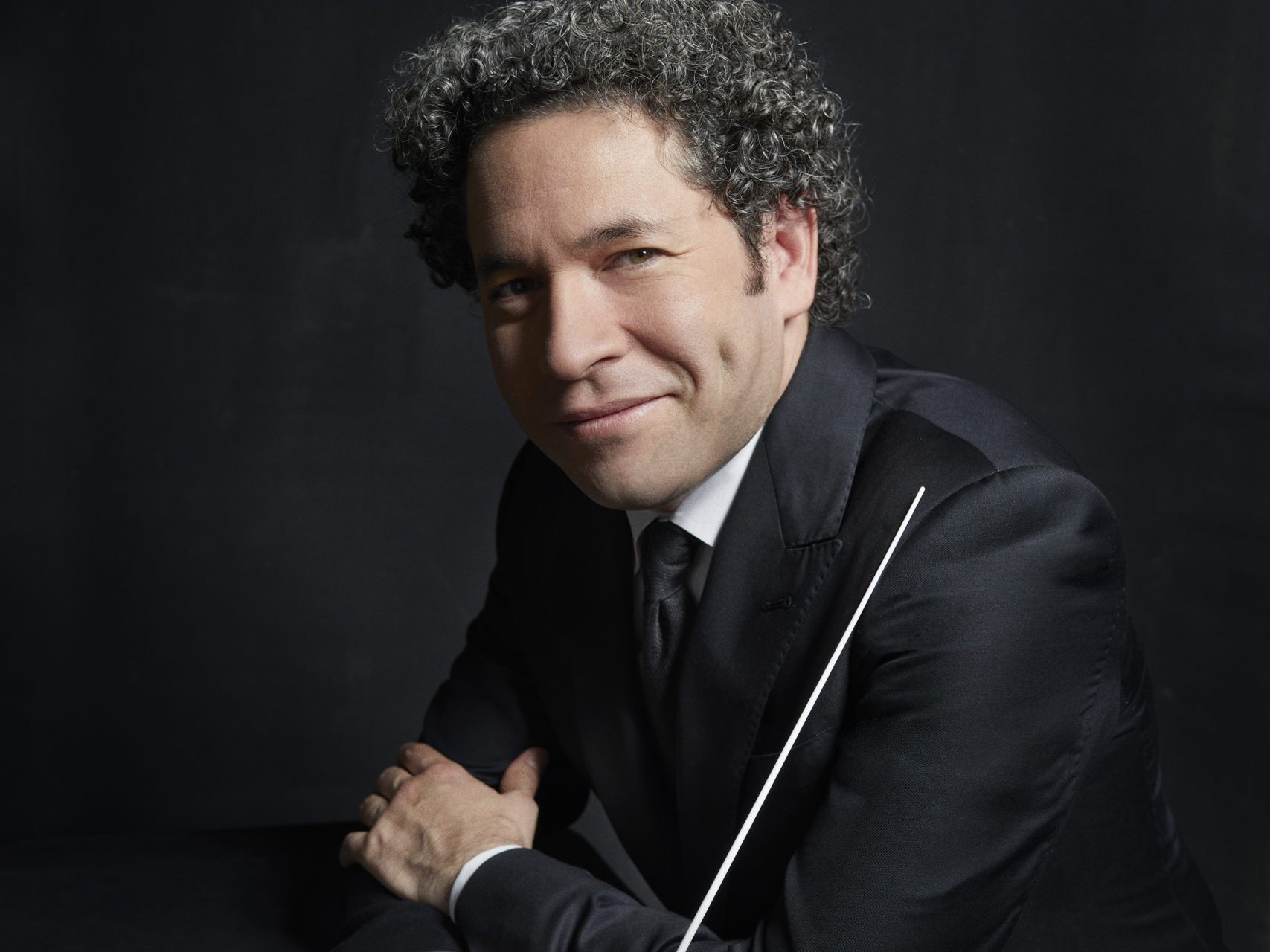 Gustavo Dudamel mit lockigem Haar im Anzug lehnt an einem dunklen Tisch und hält einen Taktstock vor einem dunklen Hintergrund. Er hat Grübchen und blickt direkt in die Kamera.