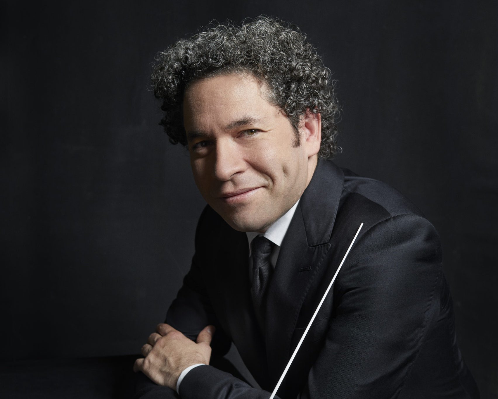 Gustavo Dudamel mit lockigem Haar im Anzug lehnt an einem dunklen Tisch und hält einen Taktstock vor einem dunklen Hintergrund. Er hat Grübchen und blickt direkt in die Kamera.