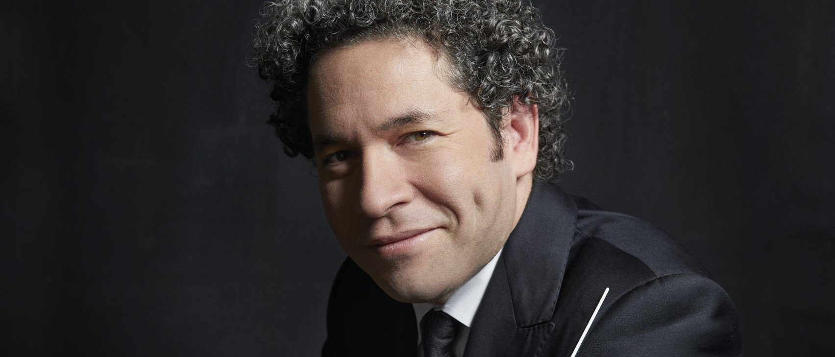 Gustavo Dudamel mit lockigem Haar im Anzug lehnt an einem dunklen Tisch und hält einen Taktstock vor einem dunklen Hintergrund. Er hat Grübchen und blickt direkt in die Kamera.