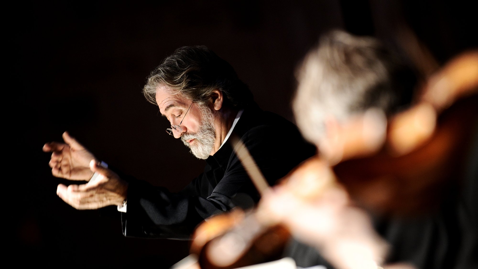 A bearded man in glasses conducts an orchestra, captured mid-motion with expressive hands. The foreground is blurred, showing musicians and string instruments out of focus.