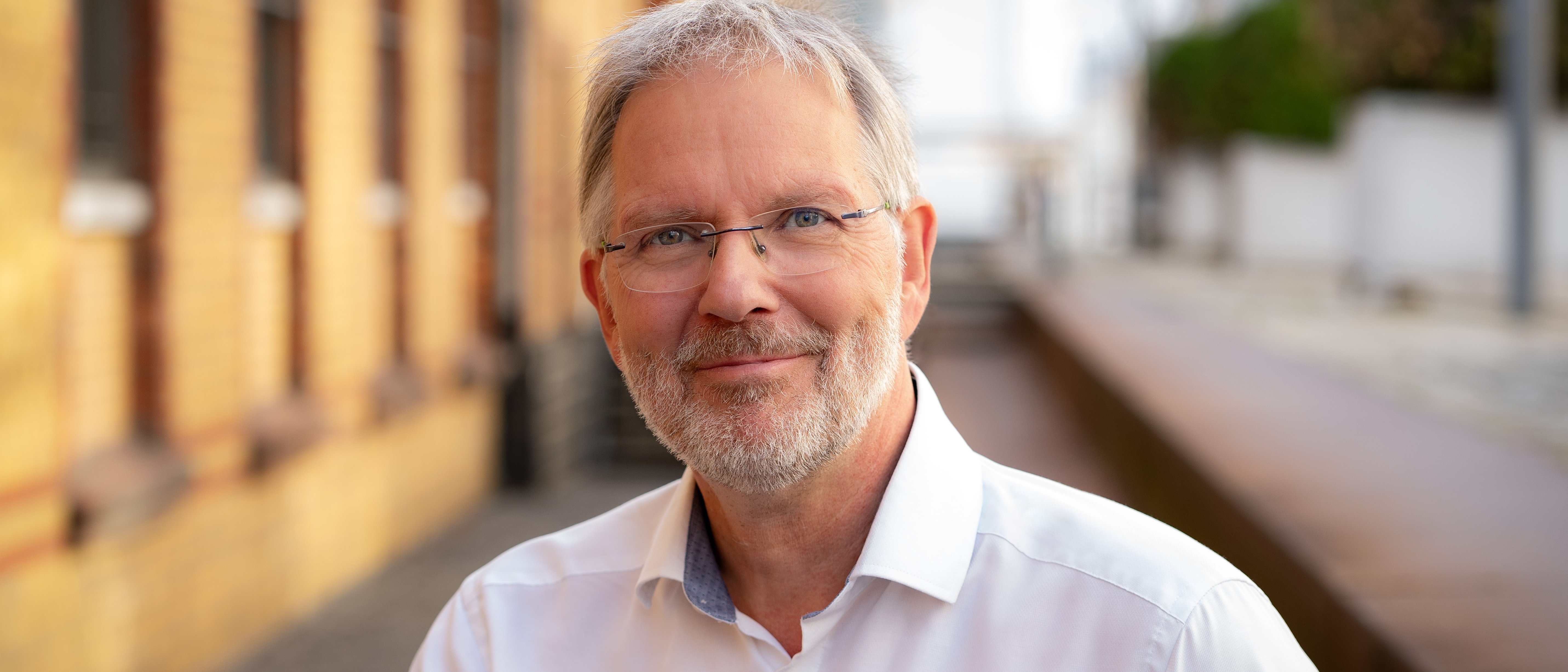 Ein Mann mit grauem Haar, Brille und Bart lächelt in die Kamera. Er trägt ein weißes Hemd und steht im Freien neben einem Backsteingebäude.