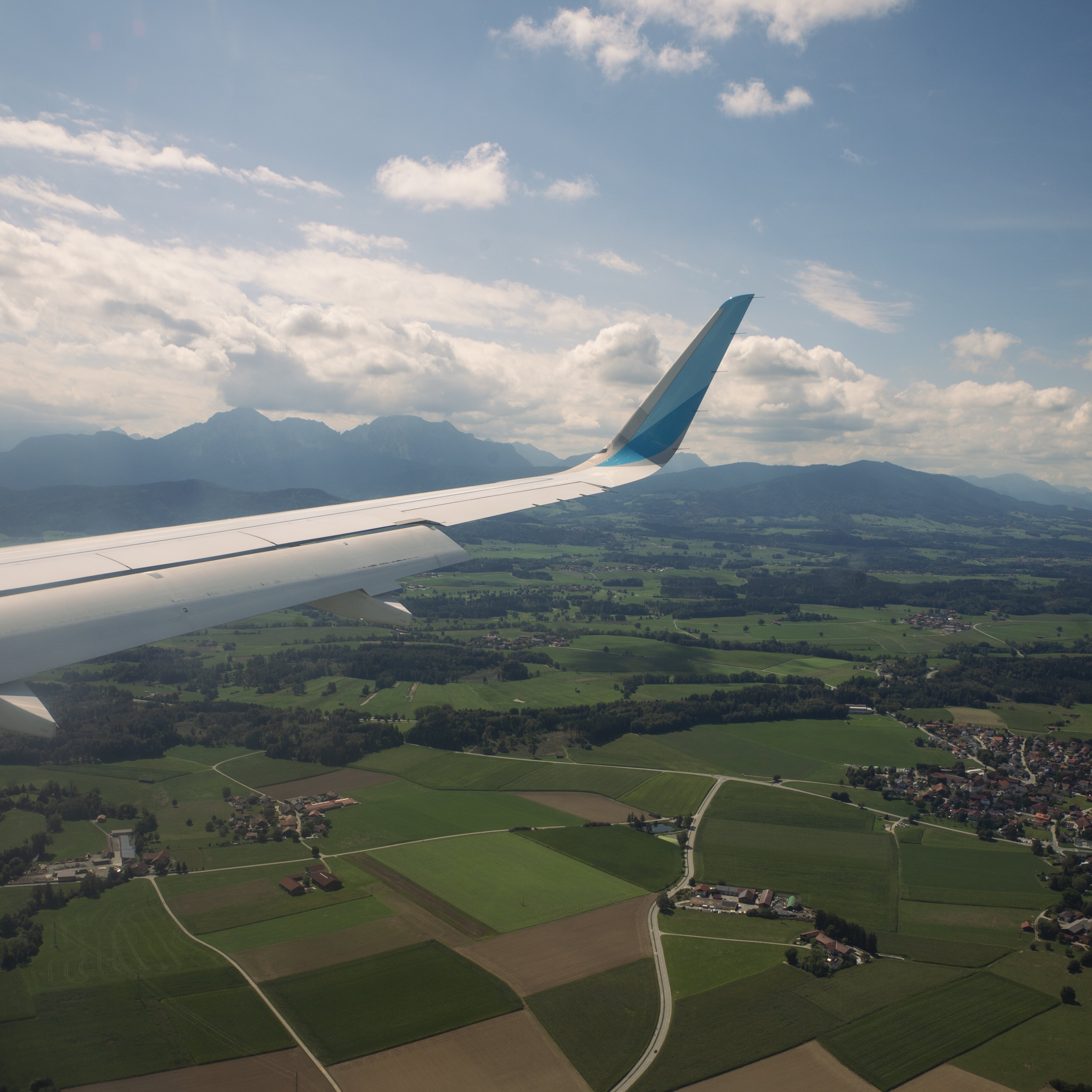 Luftaufnahme aus einem Flugzeugfenster, die einen Flügel über grünen Feldern und Ackerland zeigt, mit vereinzelten Häusern, kurvenreichen Straßen und Bergen in der Ferne unter einem teilweise bewölkten Himmel.