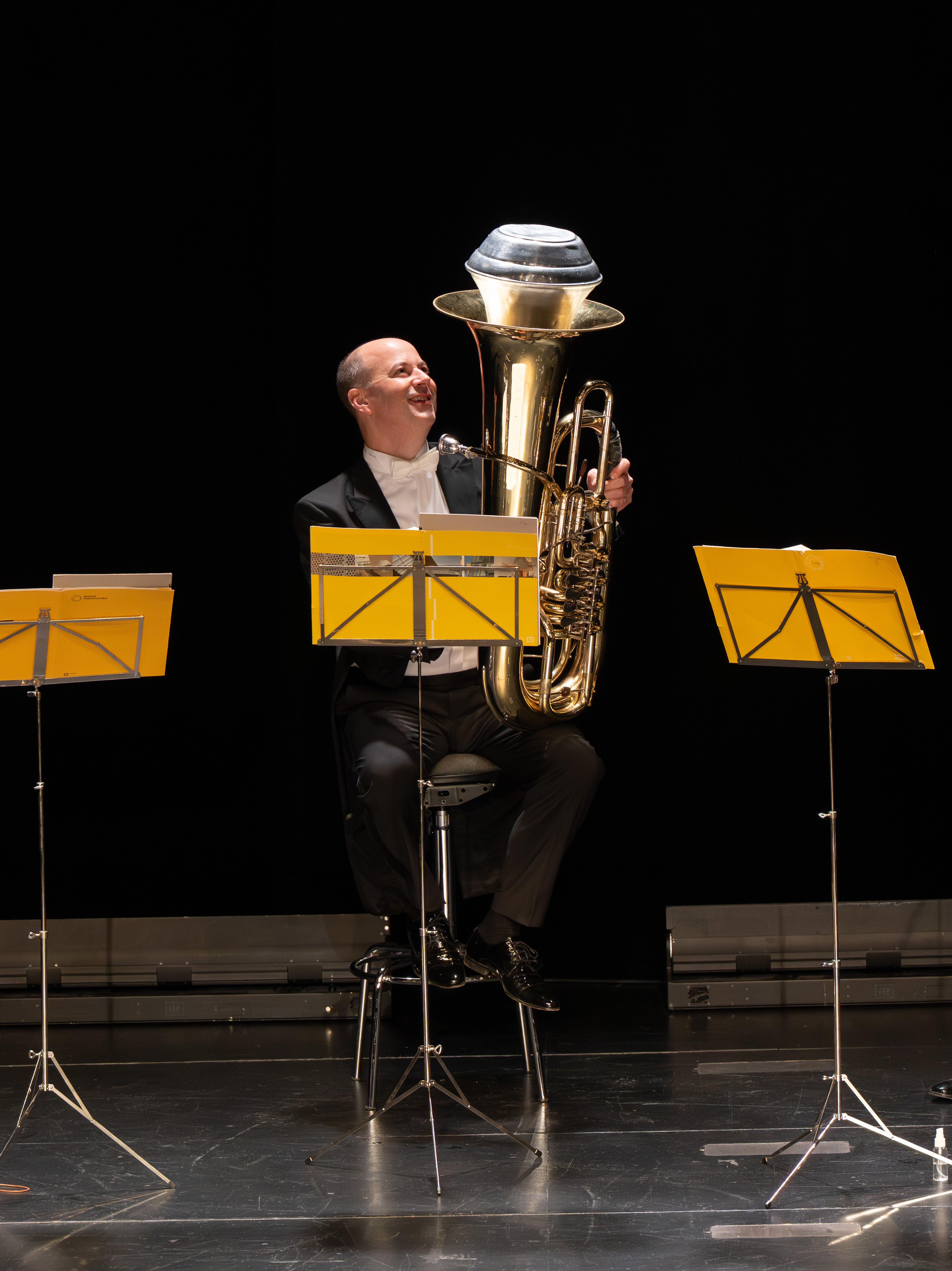 Ein Mann in formeller Kleidung sitzt auf einem Hocker, lächelt und hält eine große Messingtuba in der Hand, vor ihm auf einer dunklen Bühne stehen gelbe Notenständer.