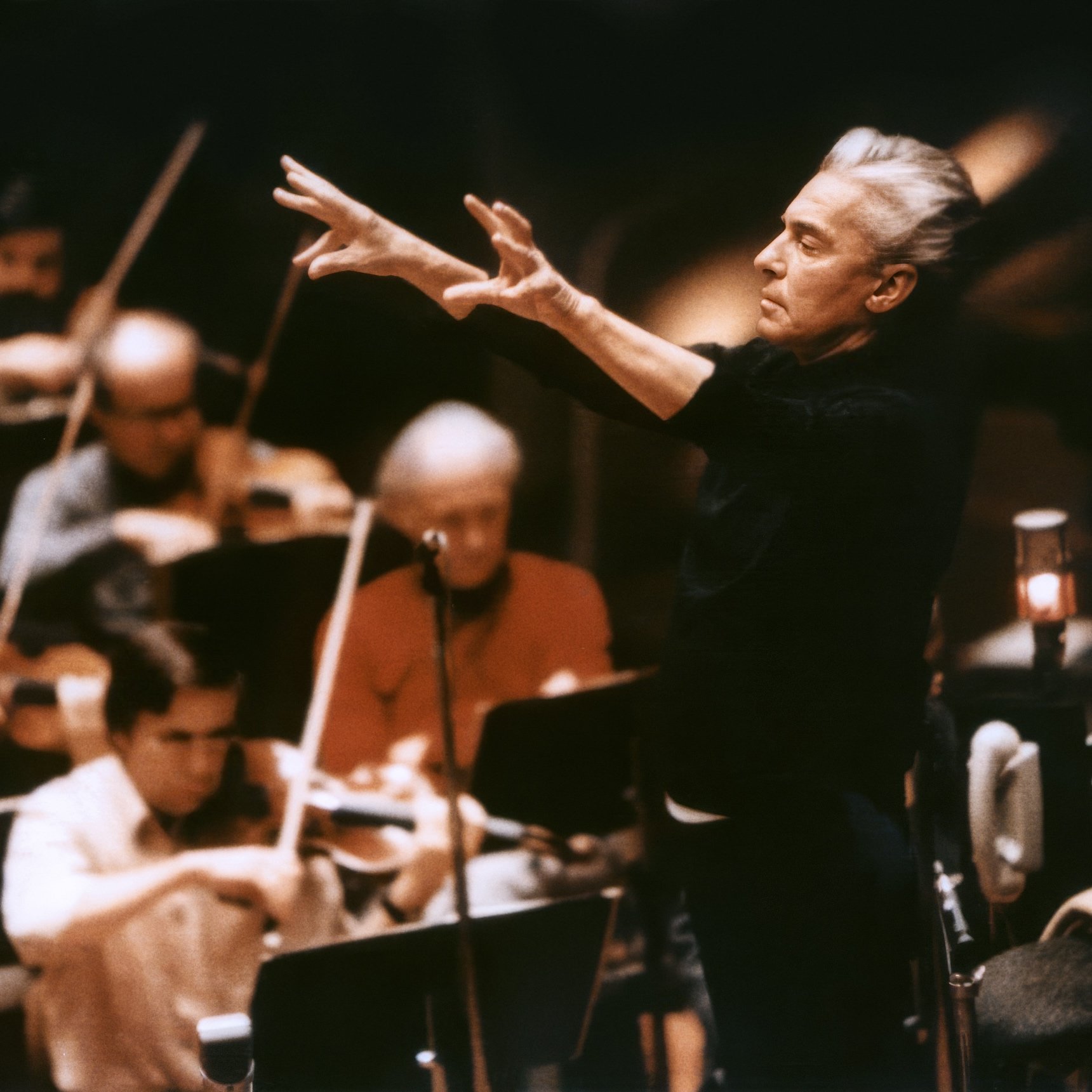 Herbert von Karajan steht mit erhobenen Armen und leitet leidenschaftlich die Berliner Philharmoniker, die in einem schwach beleuchteten Konzertsaal Streichinstrumente spielen.