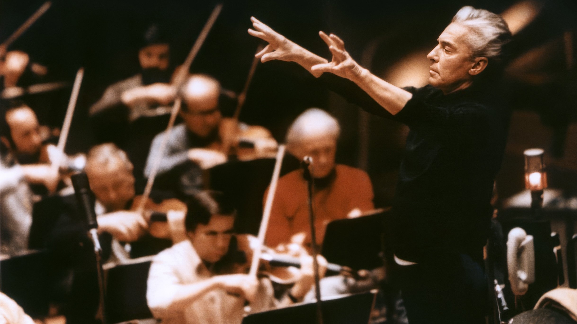 Herbert von Karajan steht mit erhobenen Armen und leitet leidenschaftlich die Berliner Philharmoniker, die in einem schwach beleuchteten Konzertsaal Streichinstrumente spielen.