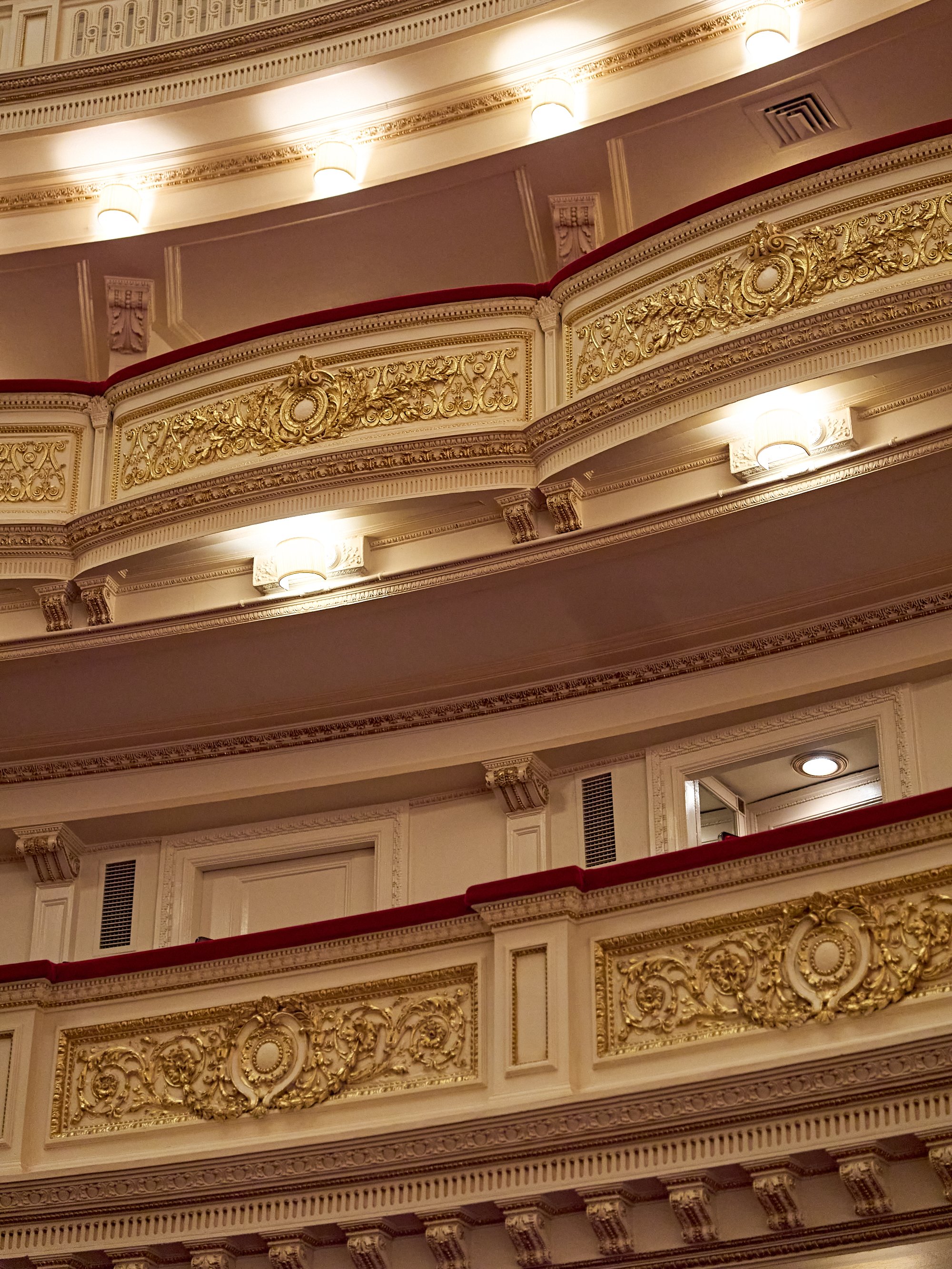 Stufenförmig angeordnete Balkone eines kunstvoll gestalteten Theaters mit weißen architektonischen Details, filigranen goldenen Verzierungen und Geländern aus rotem Samt.