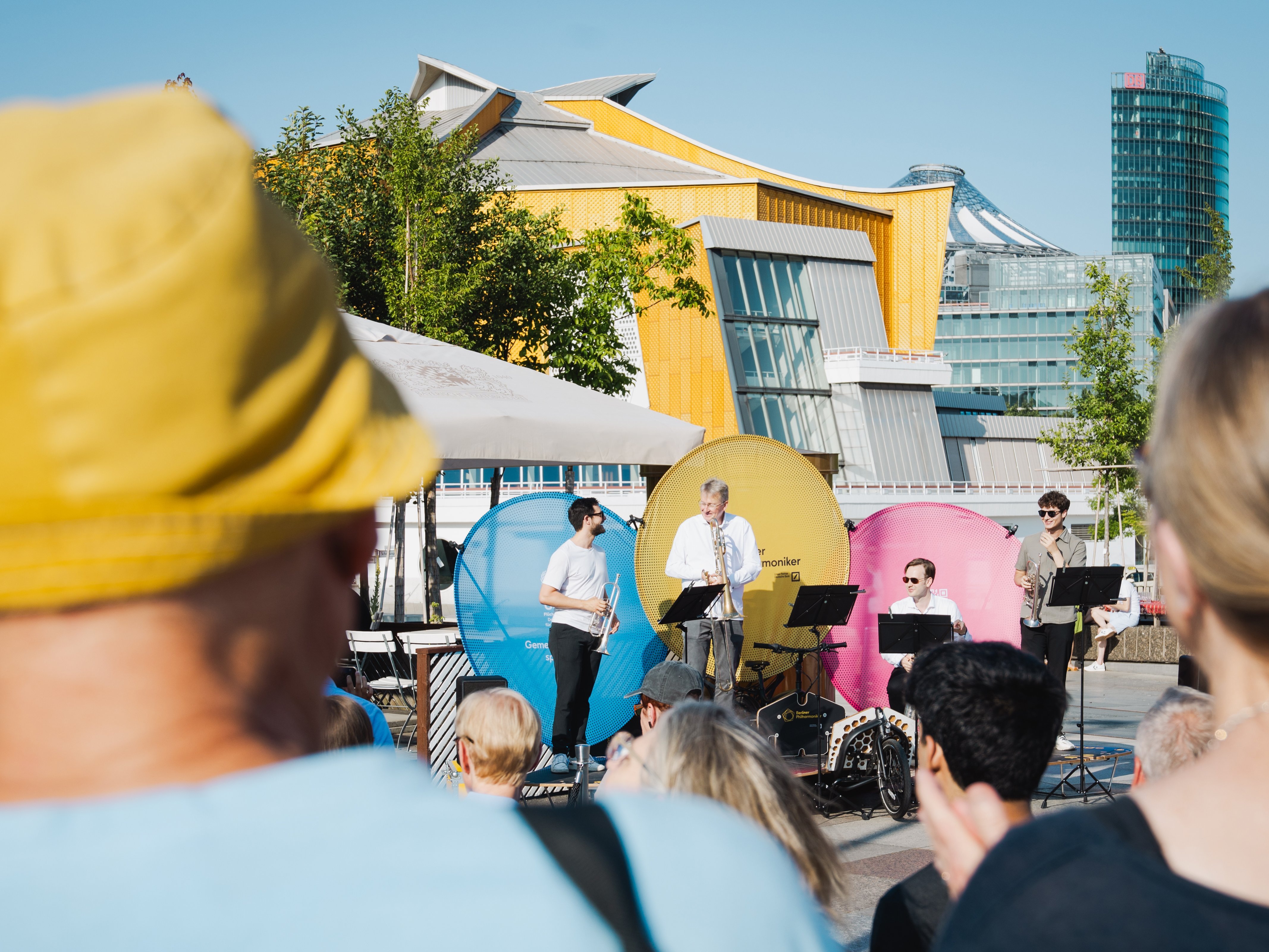 Menschen beobachten ein Konzert auf einer Fahrradbühne im Freien, bei der vier Musiker spielen; die Philharmonie Berlin und Bäume sind unter einem klaren blauen Himmel zu sehen.