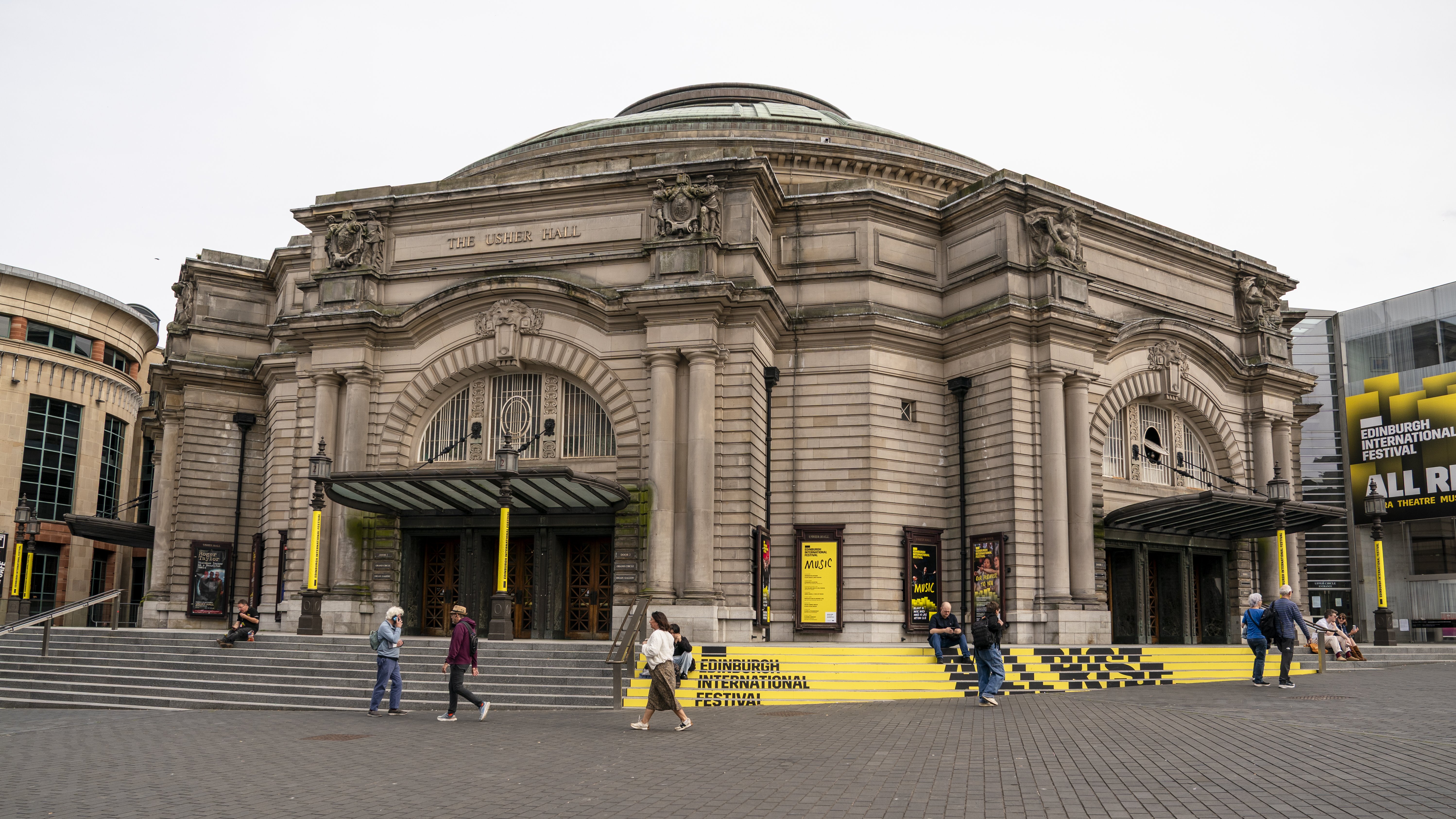 Ein großes Sandsteingebäude mit Säulen und Bögen, Menschen, die draußen vorbeigehen, und gelbe Banner mit der Aufschrift „Edinburgh International Festival» auf den Stufen und an den Türen.