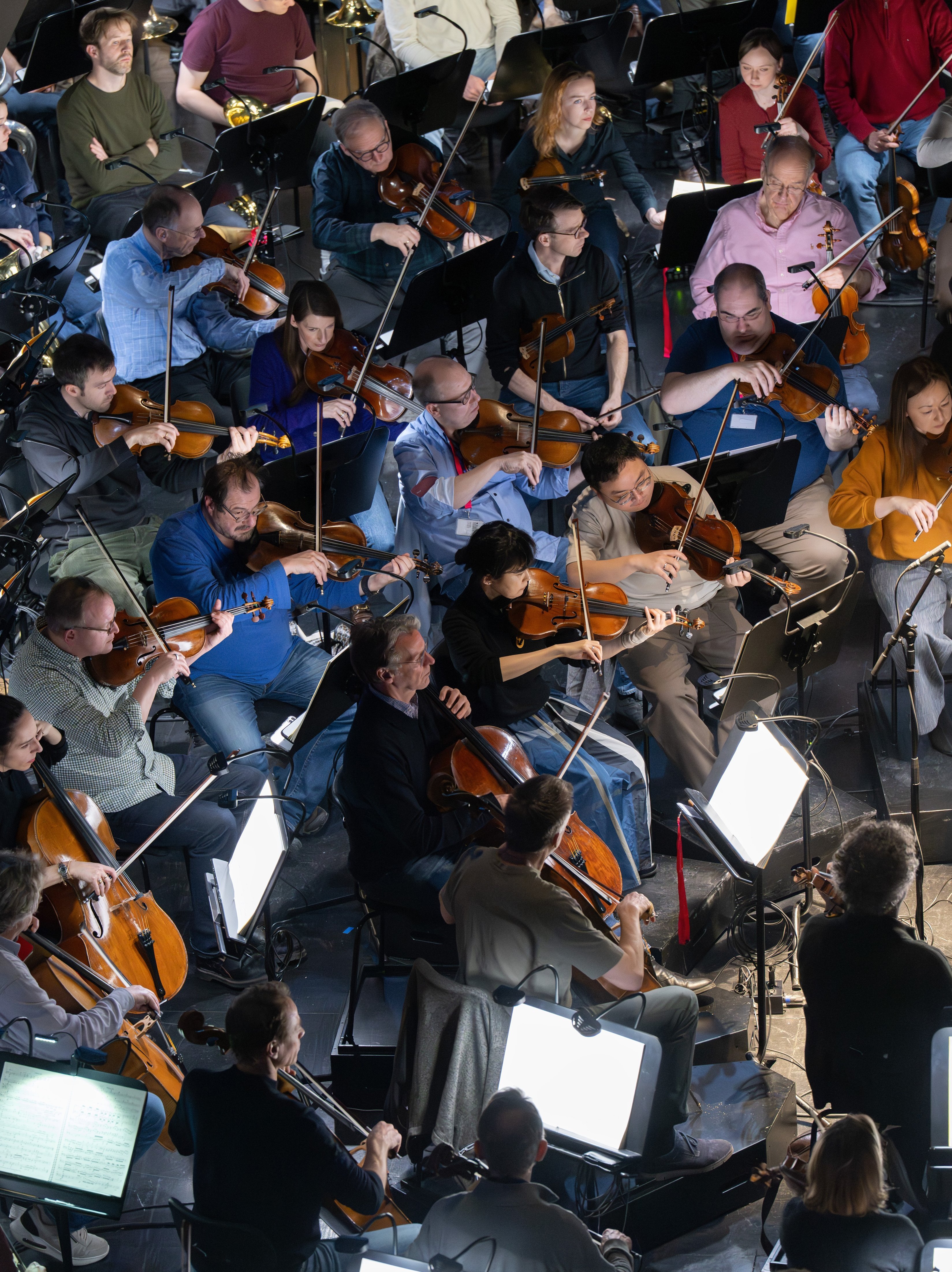 Eine Gruppe von Musikern sitzt dicht beieinander in einem Orchester und spielt Streichinstrumente wie Geigen, Bratschen und Celli. Notenblätter und Notenständer sind im hellen Scheinwerferlicht zu sehen.