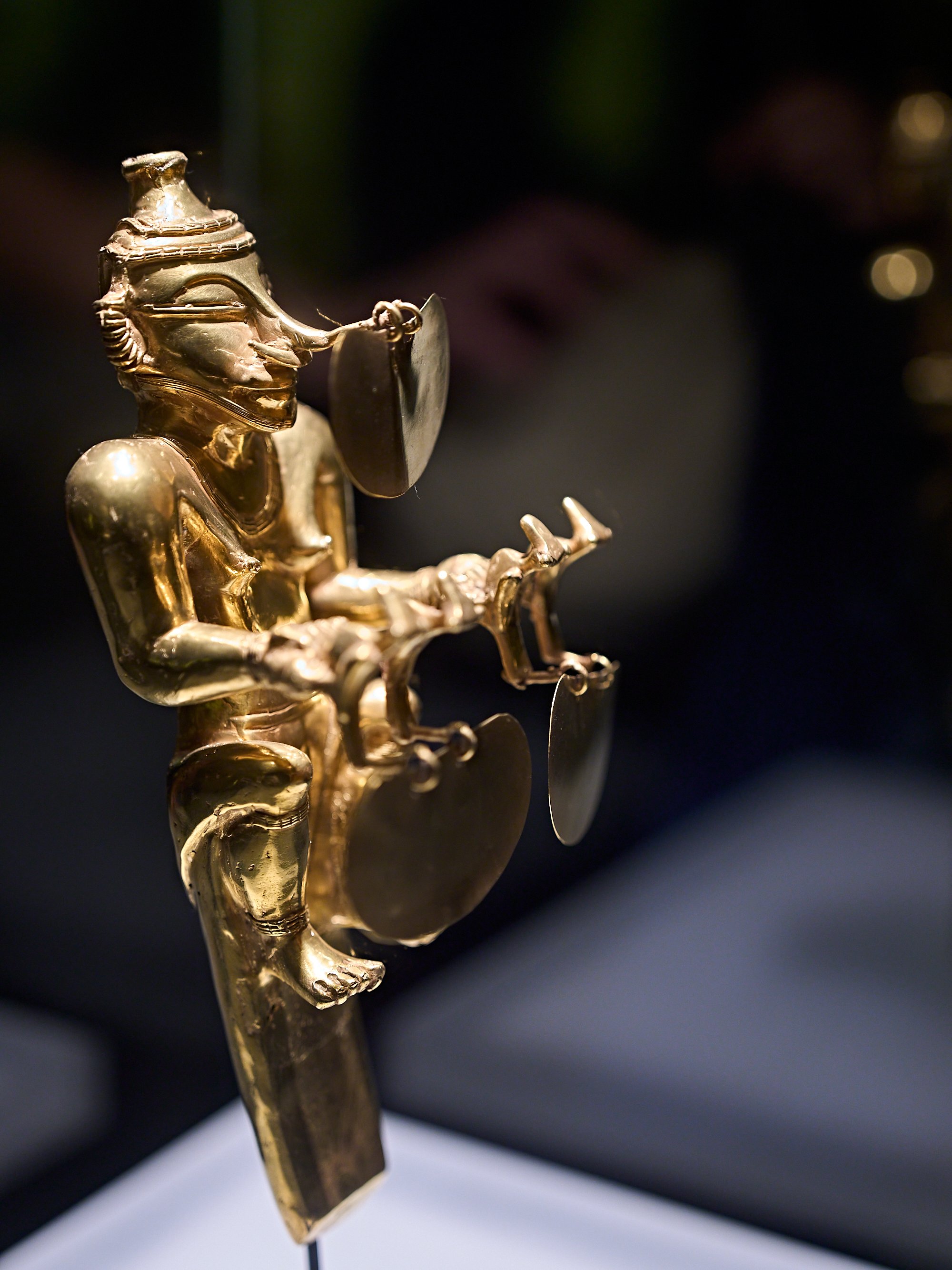 Präkolumbianische Goldfigur einer sitzenden Person mit kreisförmigen Verzierungen im Gesicht und an den Händen.