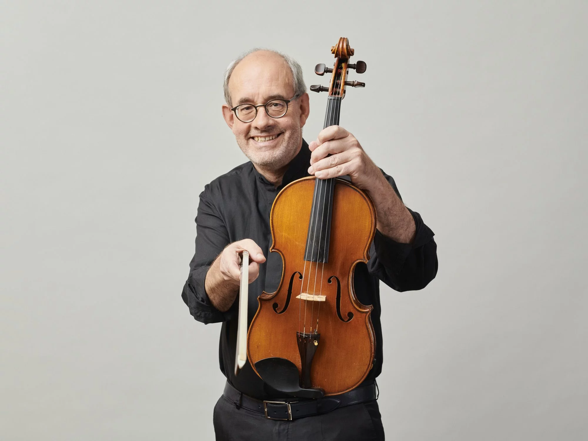 Walter Küssner mit Brille hält eine Geige und einen Bogen, trägt ein schwarzes Hemd und steht vor einem schlichten hellgrauen Hintergrund.
