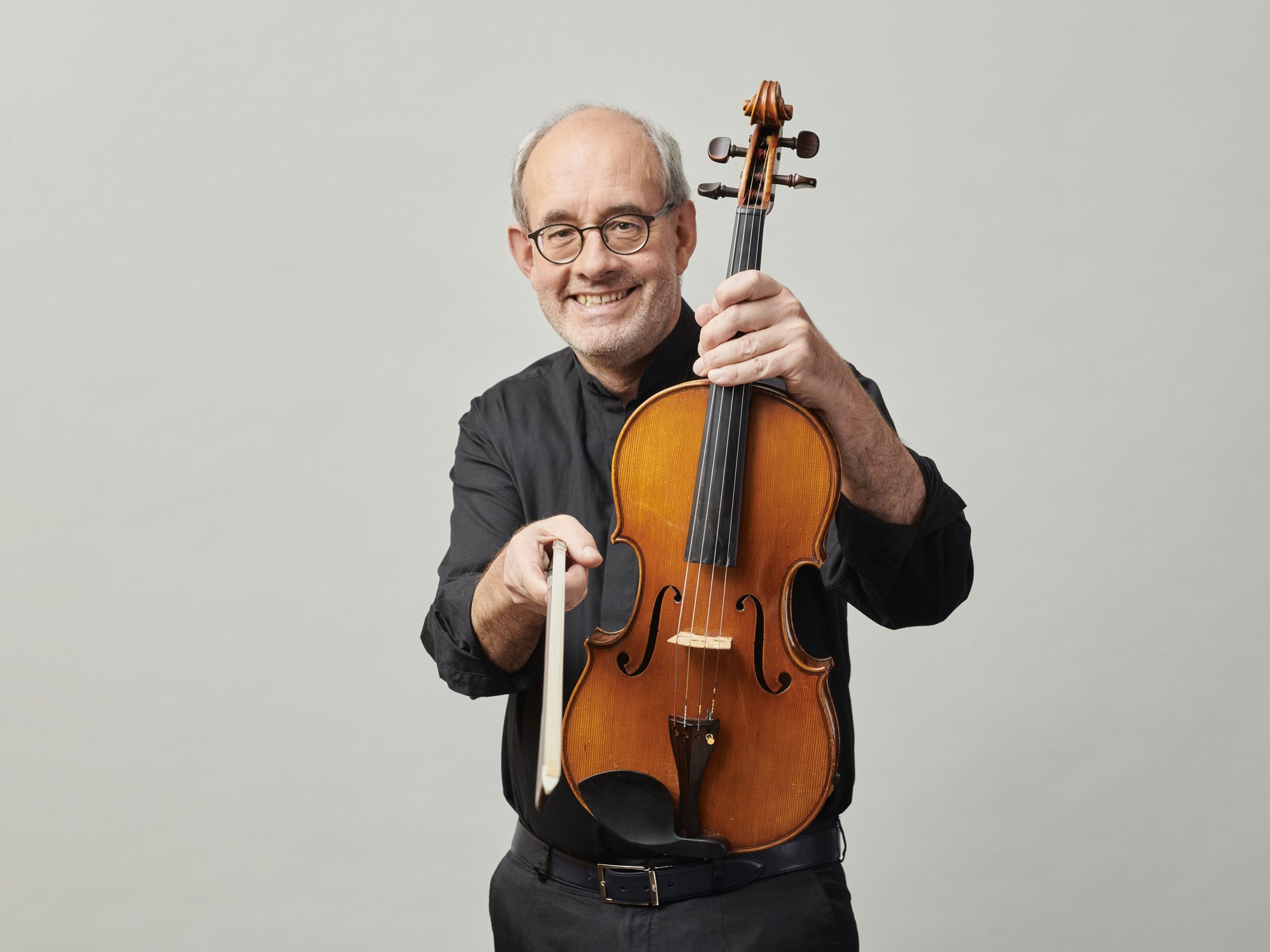 Walter Küssner, wearing glasses, holds a violin and a bow, wears a black shirt and stands in front of a plain light grey background.